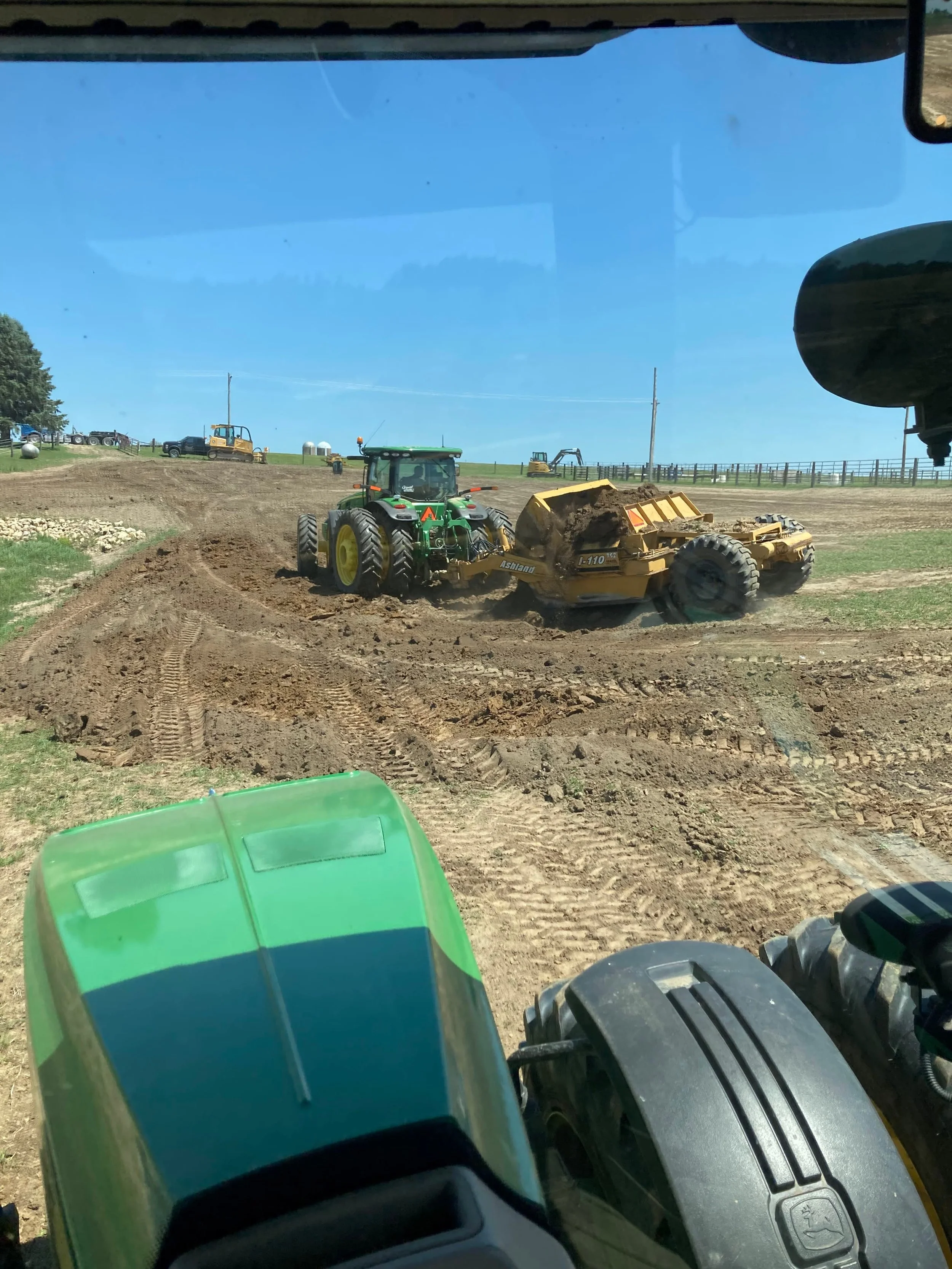 View from inside a tractor showing several tractors and construction machinery working on a farm or construction site with a clear blue sky and a fenced field in the background.