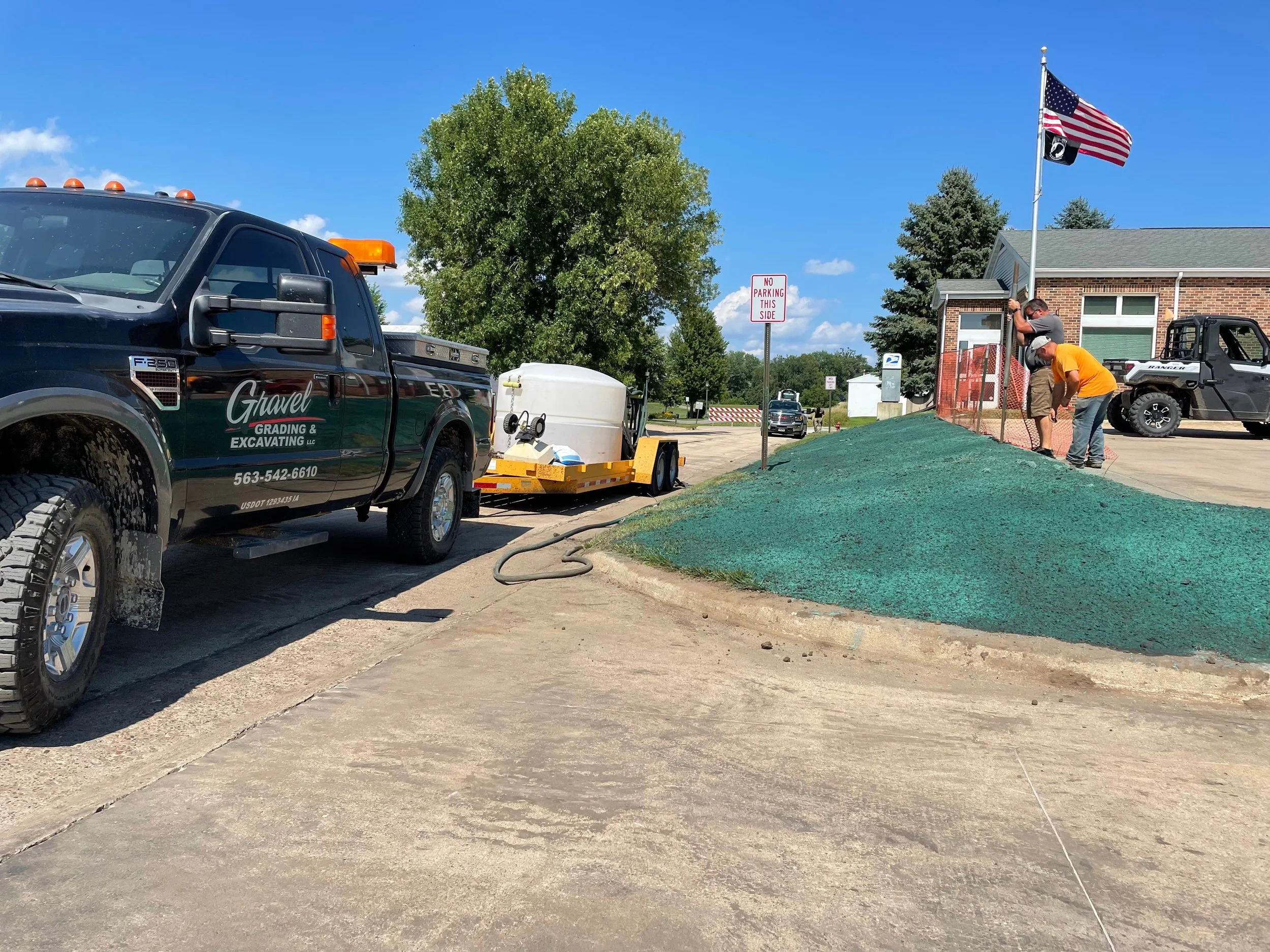 Workers installing landscape fabric on a grassy area in front of a brick building, with a flagpole flying an American flag, a tree, and a 'No Parking' sign in the background.