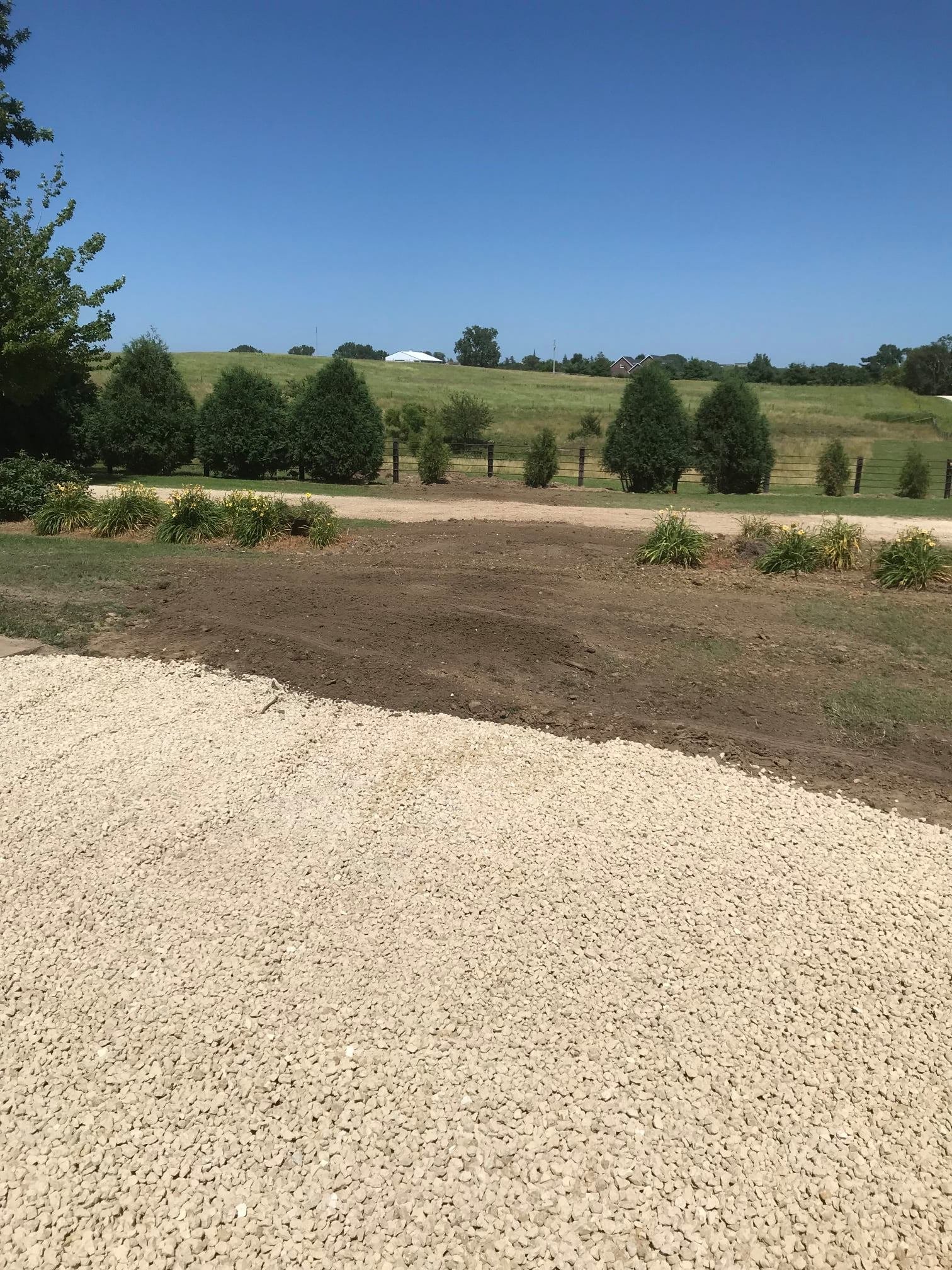 A landscape view of a yard with freshly laid gravel, some shrubs, a fenced area, rolling hills, trees, and houses in the distance under a clear blue sky.
