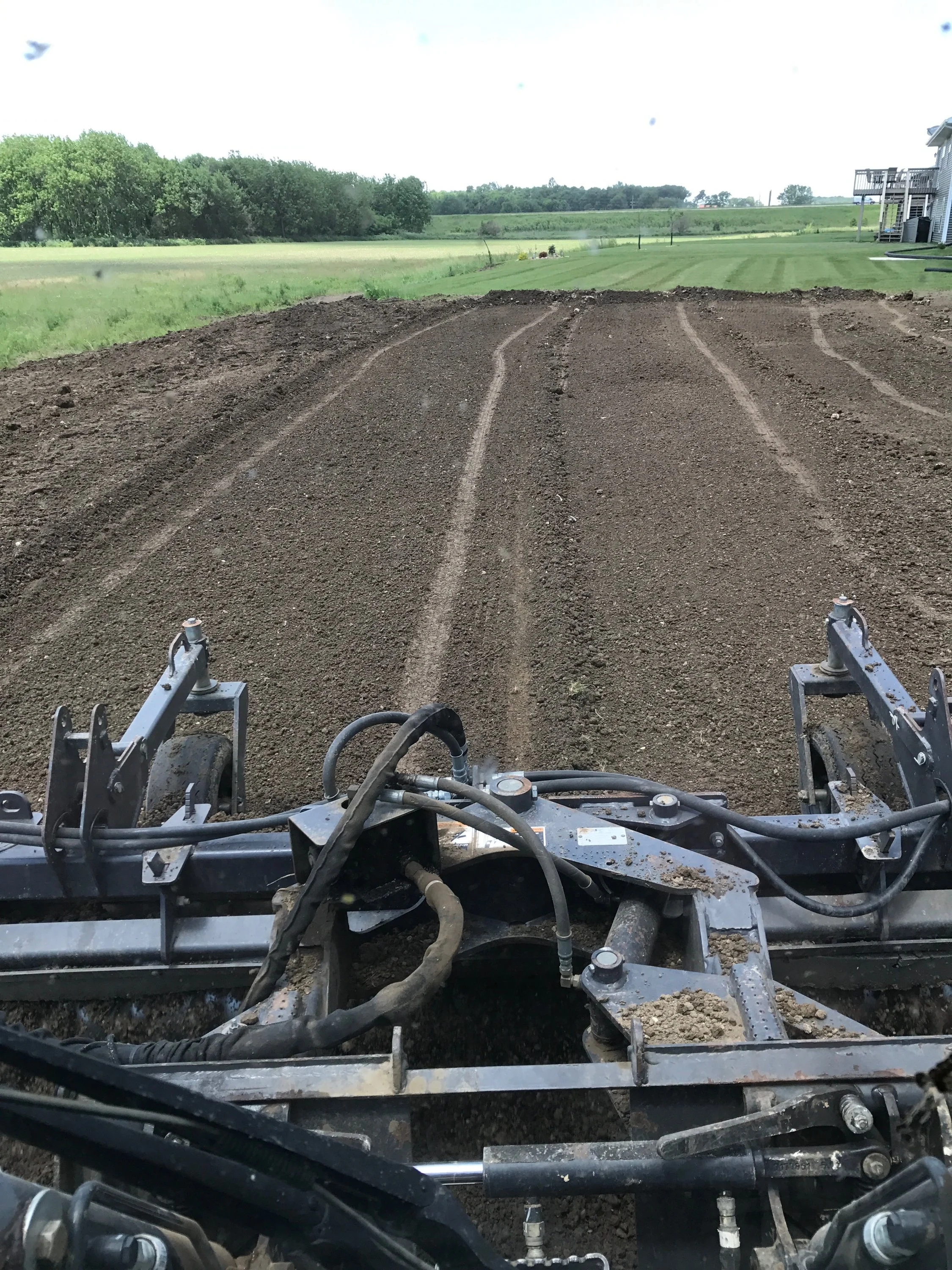 View from a skidsteer showing freshly graded topsoil in the foreground with tire tracks, green grass, a line of trees, and a house with a deck in the background.