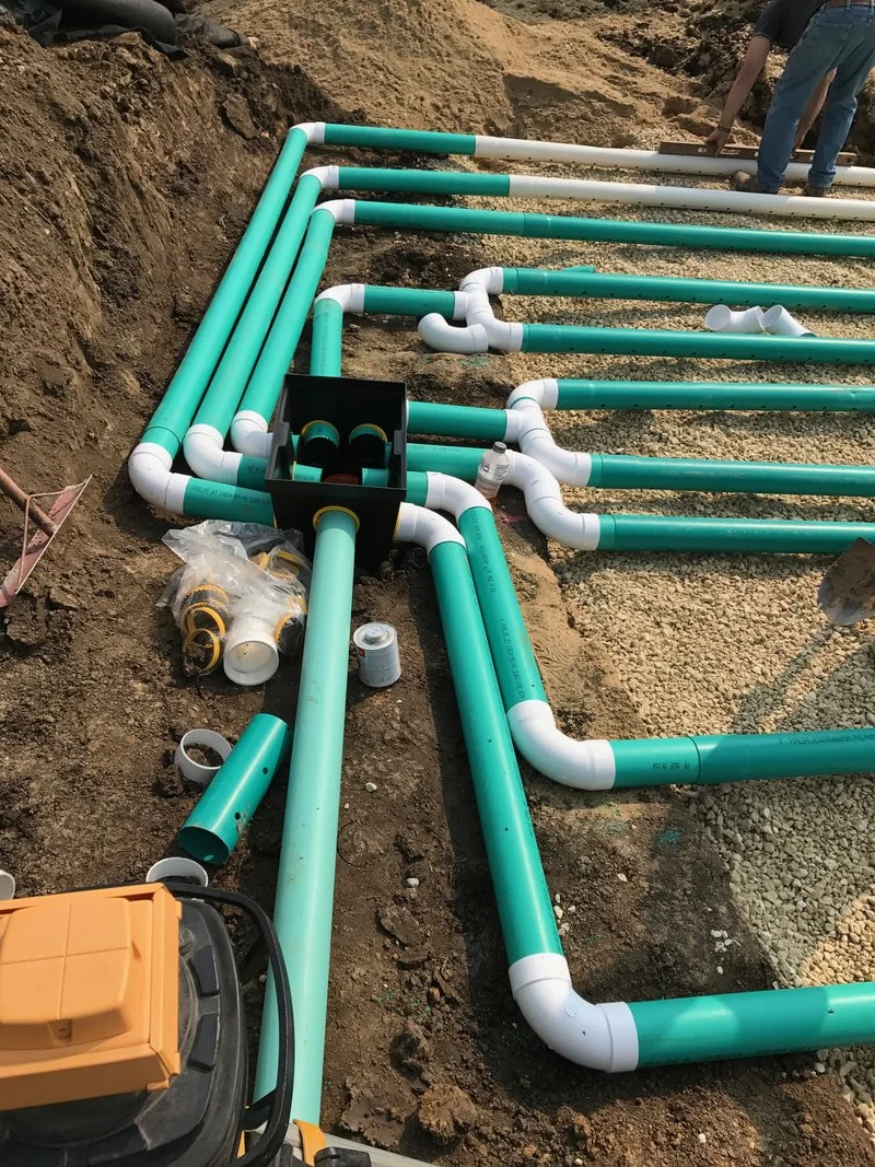 Construction site with green and white PVC pipes being installed underground, with some pipes connected and others waiting to be fitted, and a worker in jeans working nearby.