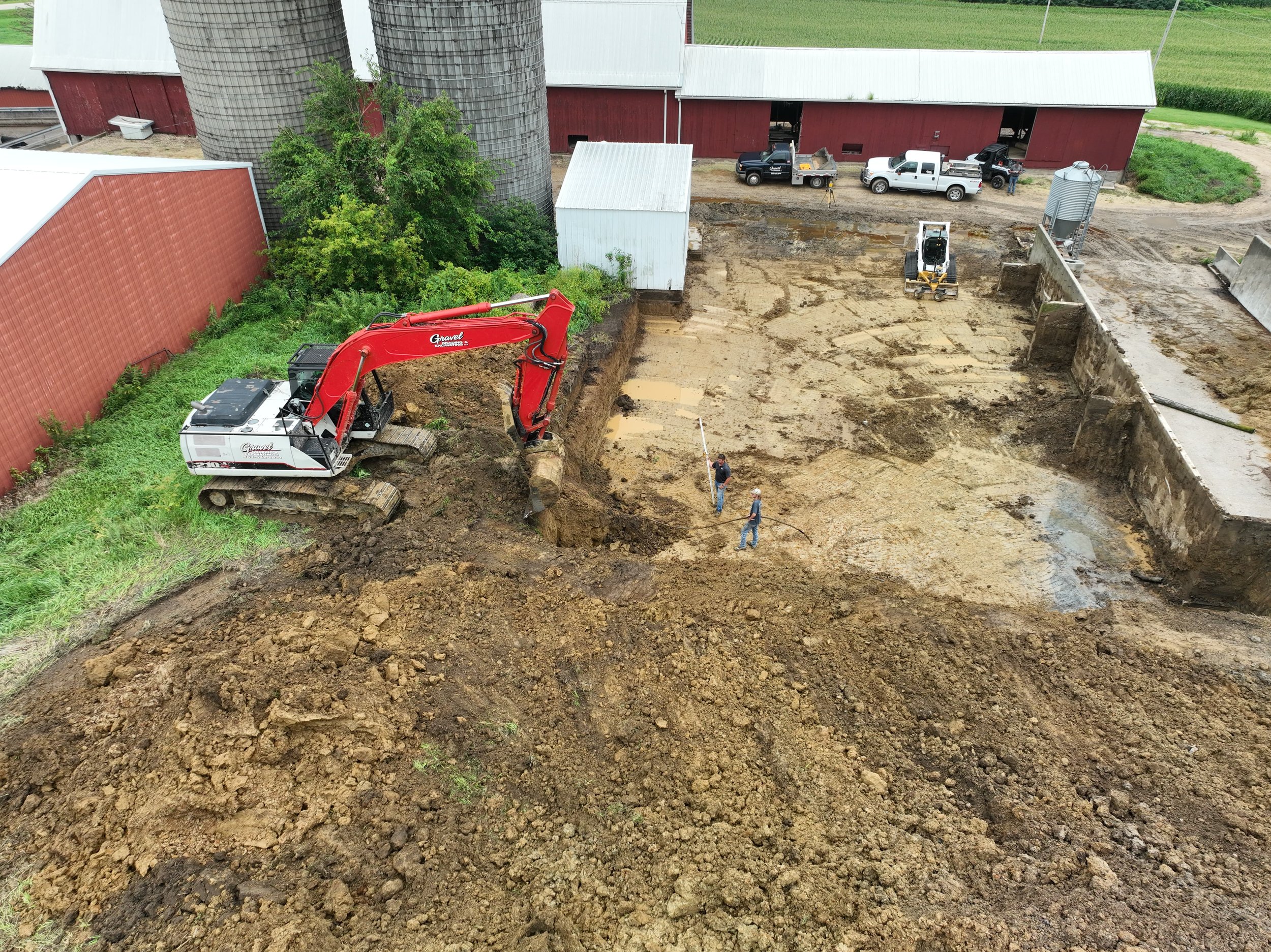 View of a construction site with excavator, two people standing and surveying, trucks parked near a red barn, and two silos in the background.