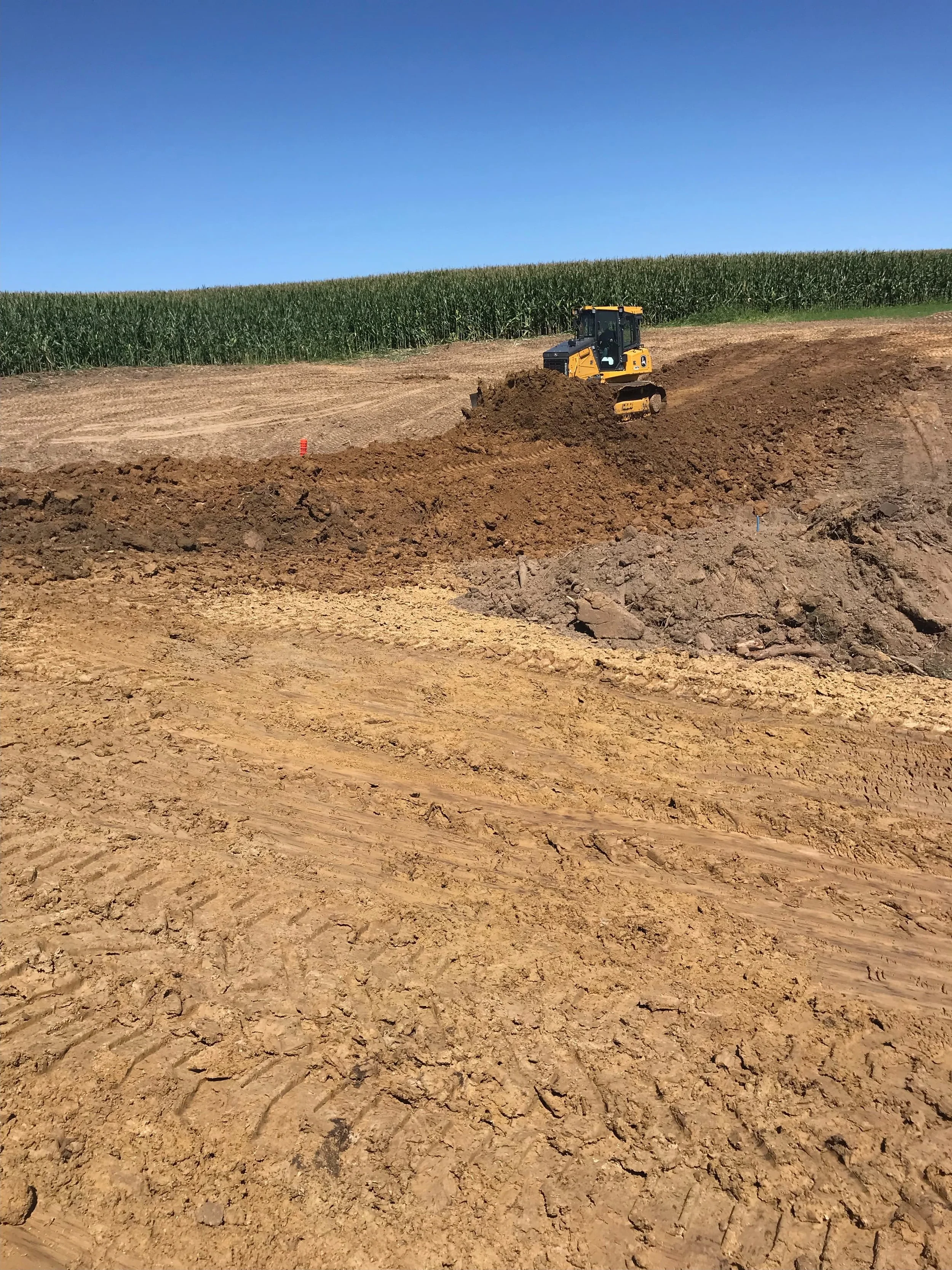 A yellow construction vehicle working on dirt in the foreground, with a field of green corn and a clear blue sky in the background.