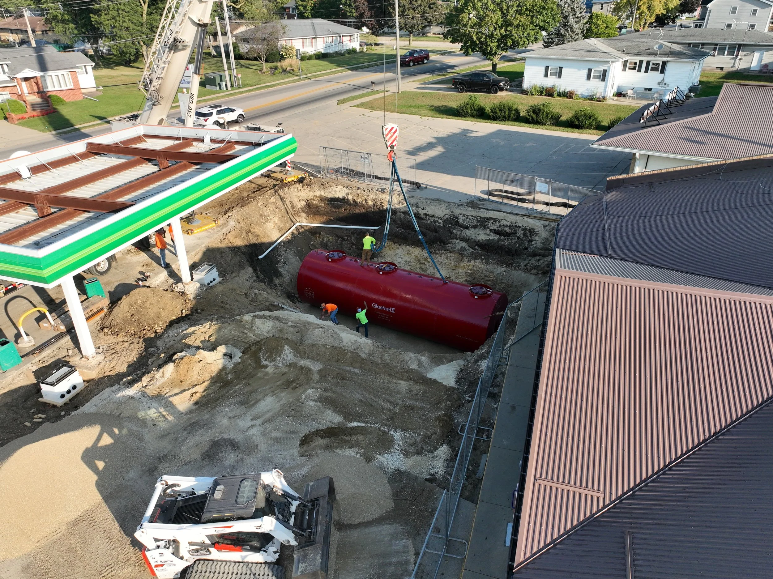 Construction workers are installing a large red tank on a construction site next to a building with a metal roof. There is a crane lifting the tank, and excavated soil is visible around the site. The background includes residential houses and a parking lot.