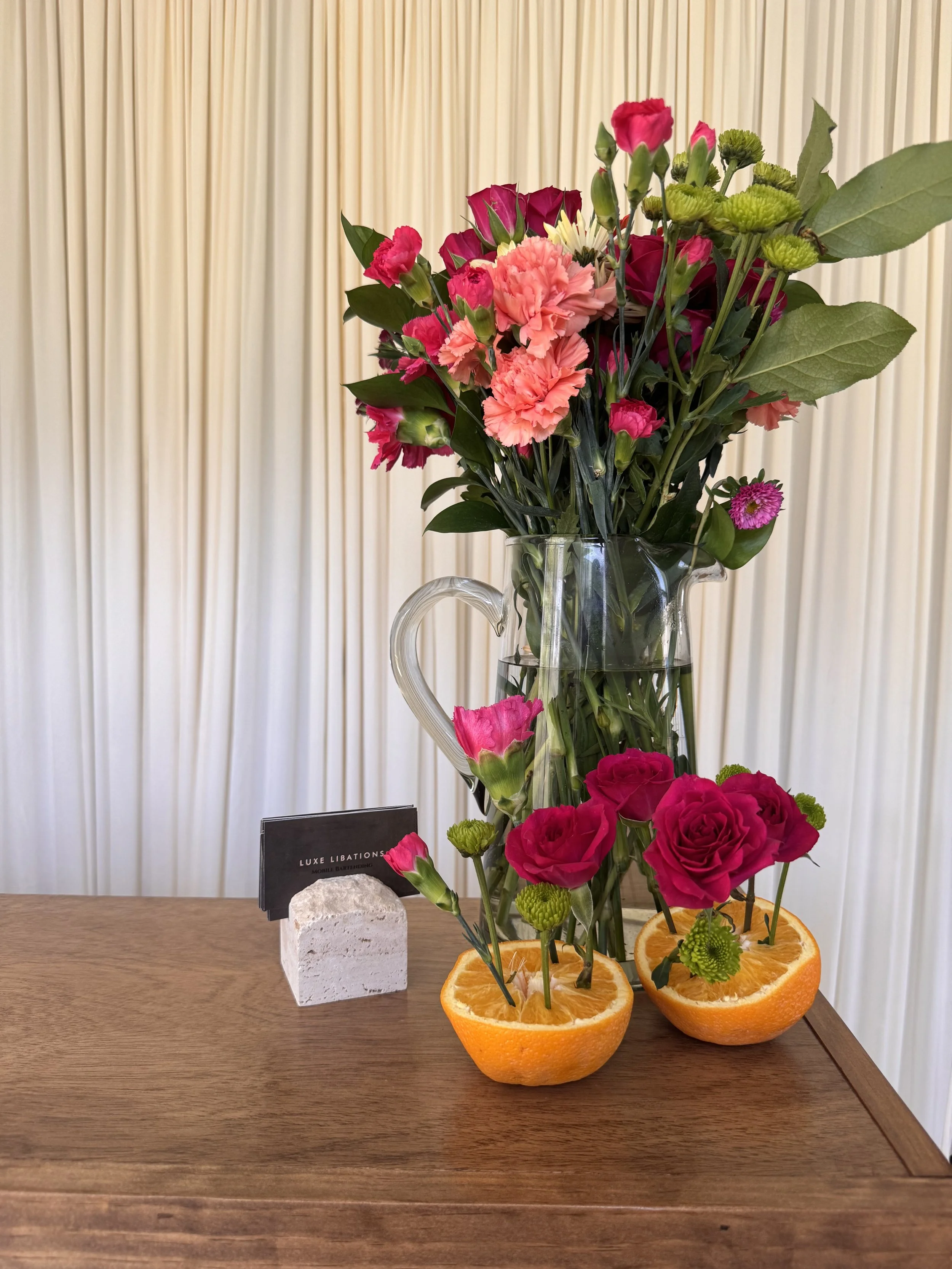 A glass vase with a bouquet of pink, red, and green flowers and leaves, placed on a wooden surface. The bouquet includes carnations, roses, and other flowers. The vase is behind two halved oranges with flowers inserted into them, and a small black card and a white stone are on the left side of the surface. The background features cream-colored vertical blinds.