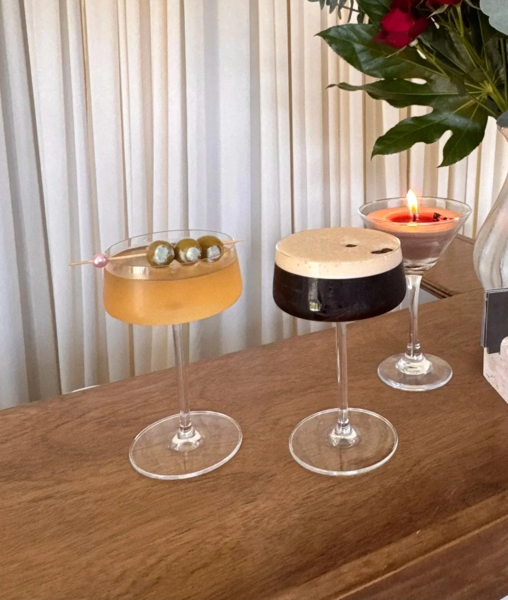 Three wine glasses with drinks on a wooden table, decorated with a candle, a potted poinsettia plant and a small bowl of olives, with a white striped wall in the background.