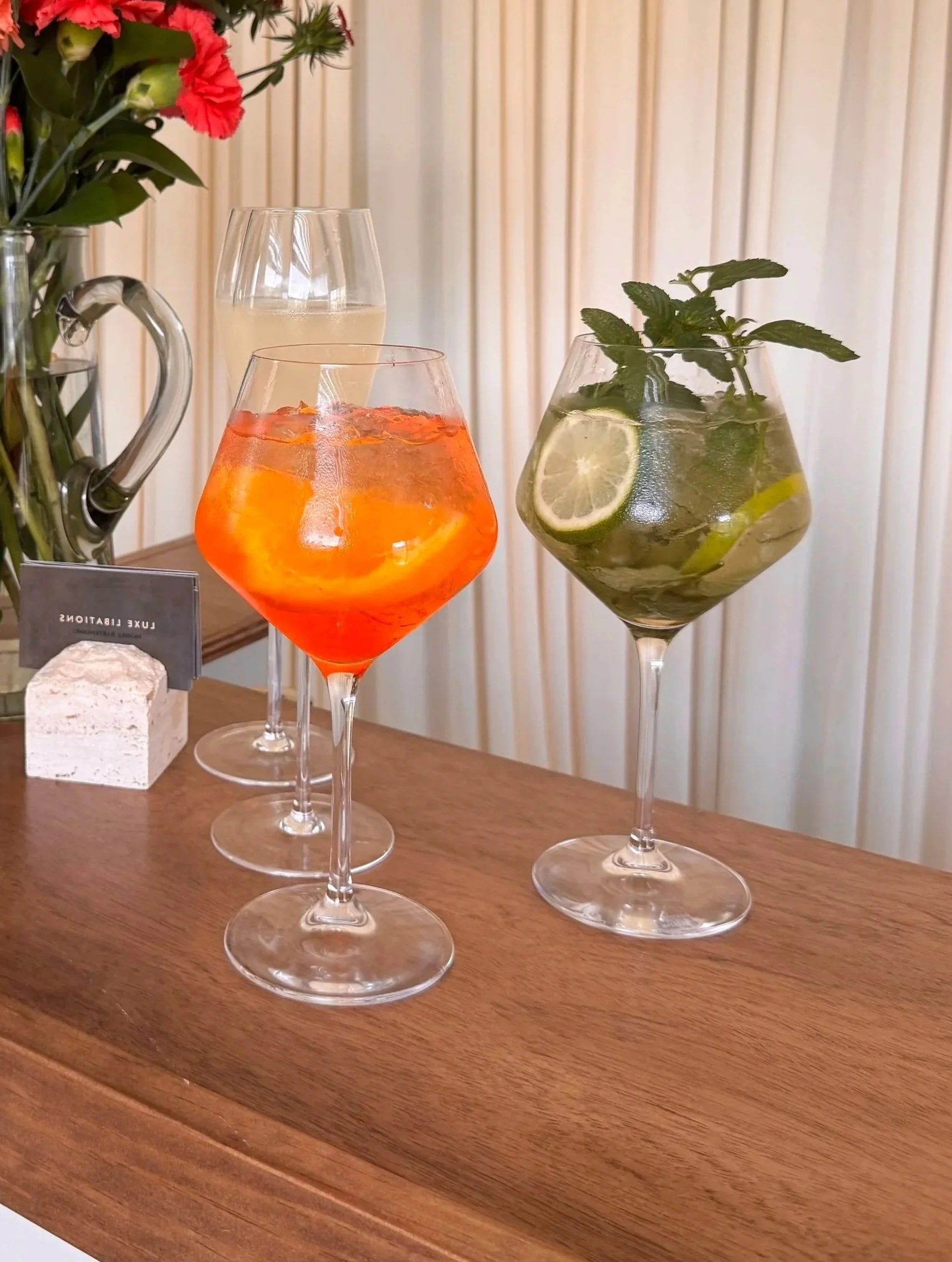Three cocktail glasses with drinks, one with orange-red liquid, another with lemon-lime slices and mint, and the third with a clear liquid and a lemon slice. A vase with red and white flowers is in the background.