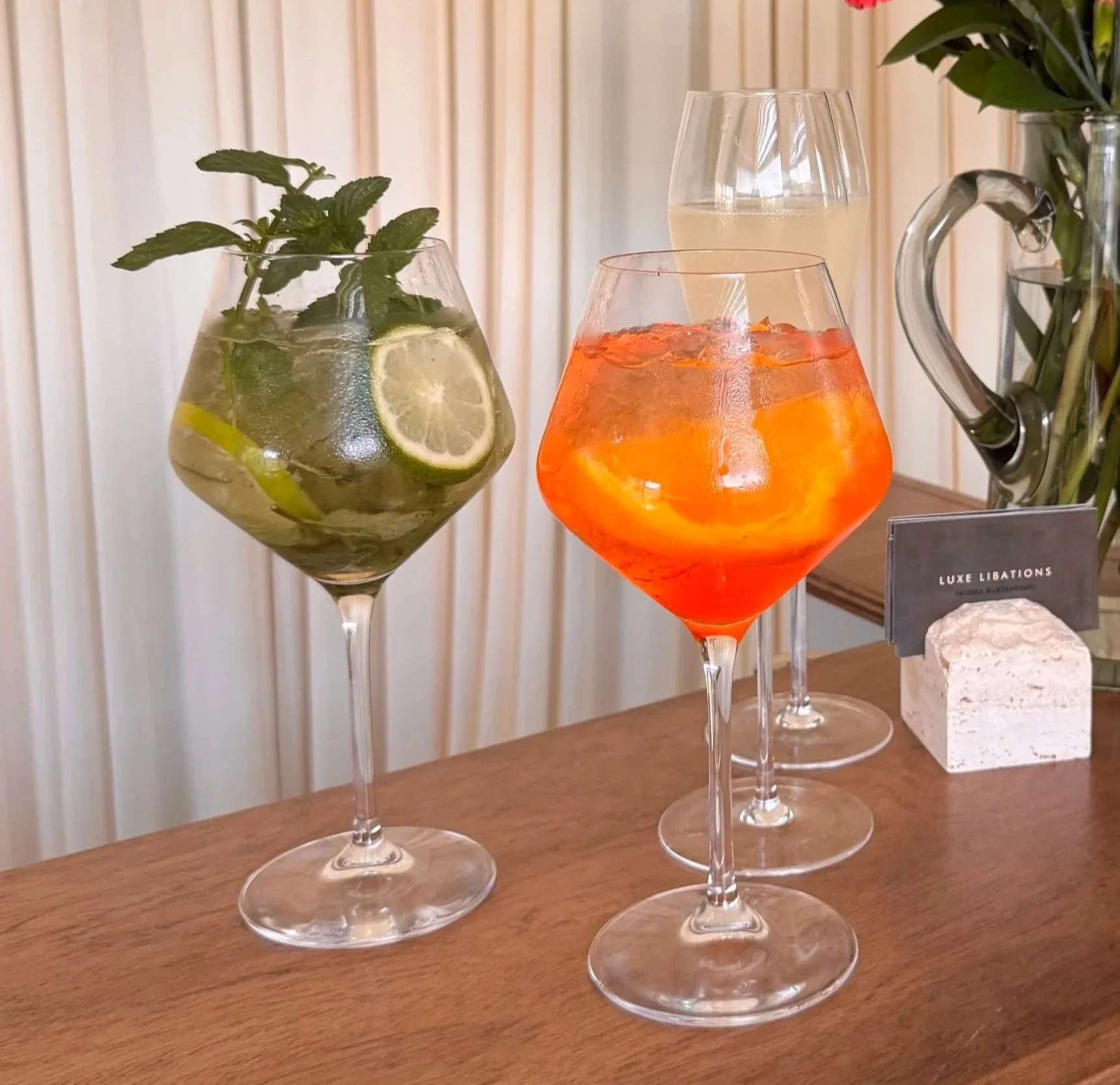 Four cocktail glasses on a wooden surface, containing colorful beverages with fruit garnishes and fresh herbs, and a vase with flowers in the background.