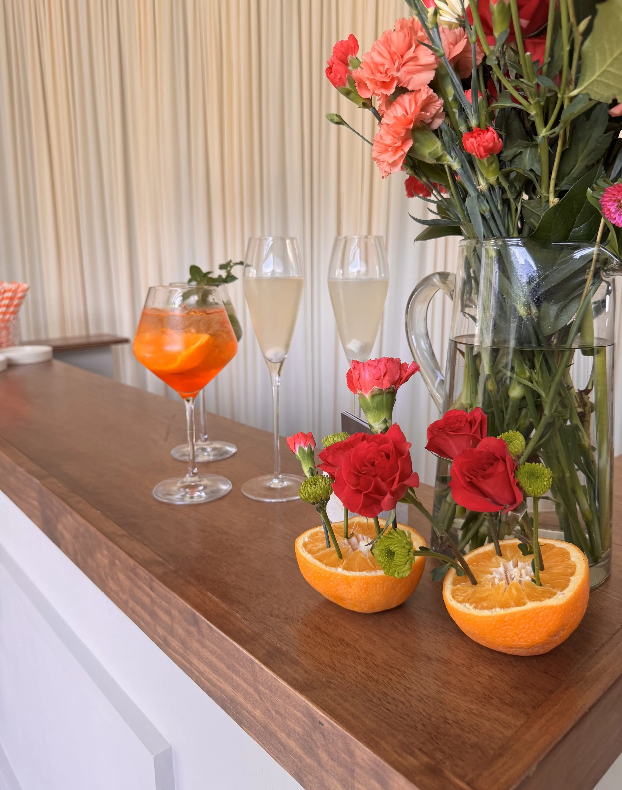 A table with a large glass vase filled with pink and red carnations, some of which are placed inside halved oranges, and a few other glasses with drinks. A smaller glass contains a drink with orange slices and ice, and some other glasses are filled with a light-colored beverage.