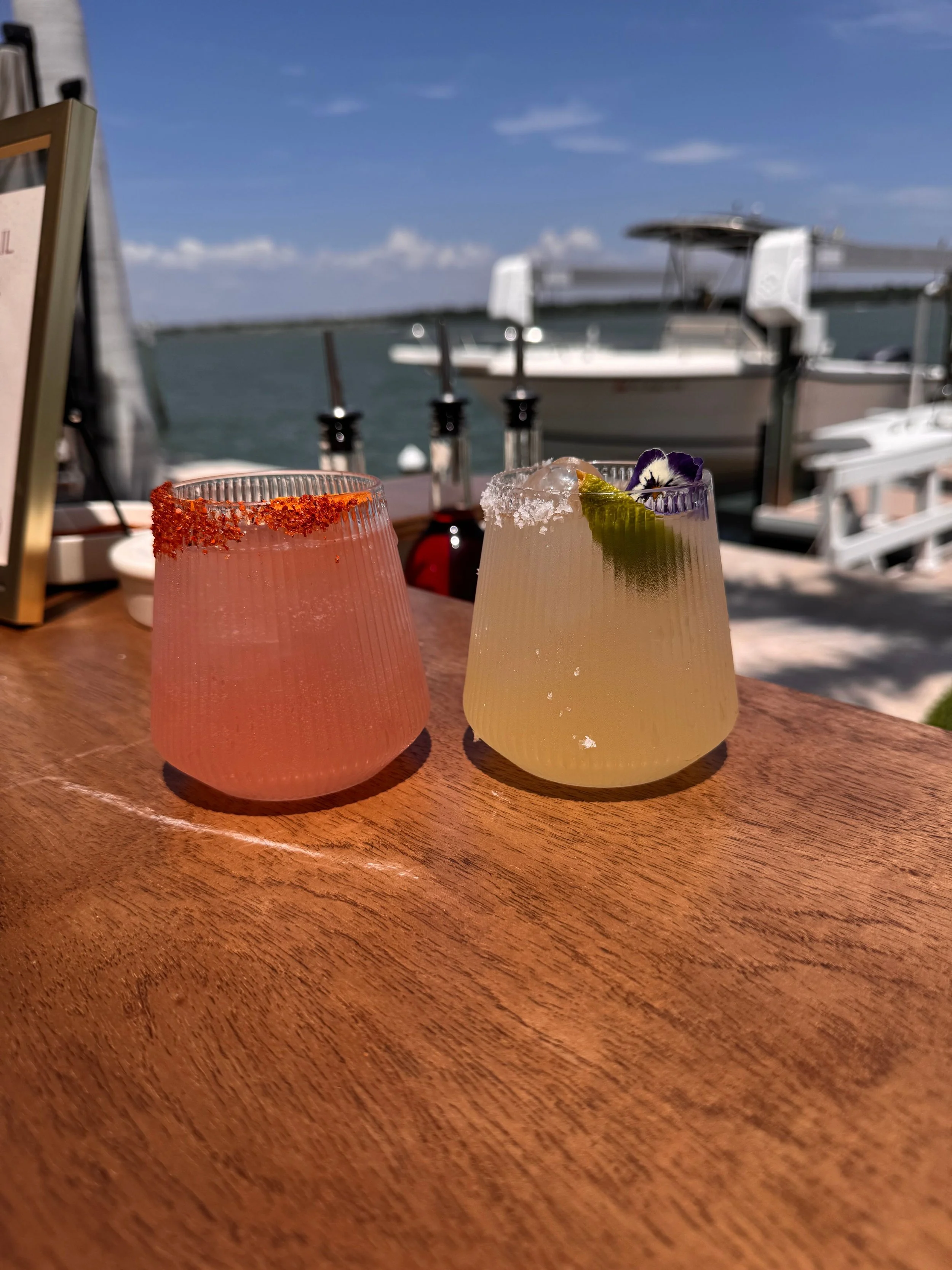 Two cocktails on a wooden table with a marina and boats in the background under blue sky.