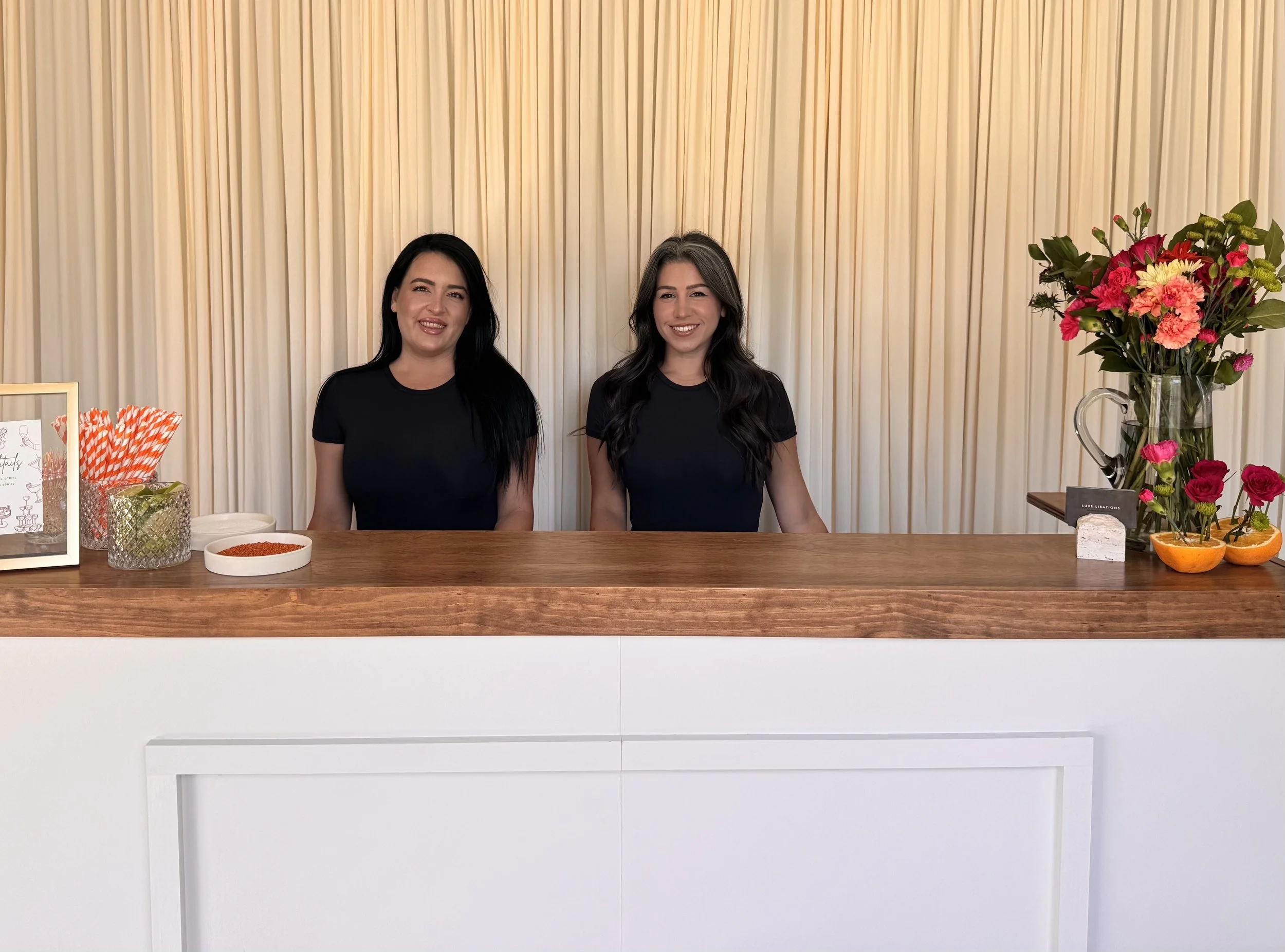 CEO & Founder, Alexis and Meghan, standing behind a white counter at an event, with a floral arrangement on the right and various small items on the left, against a beige curtain backdrop.