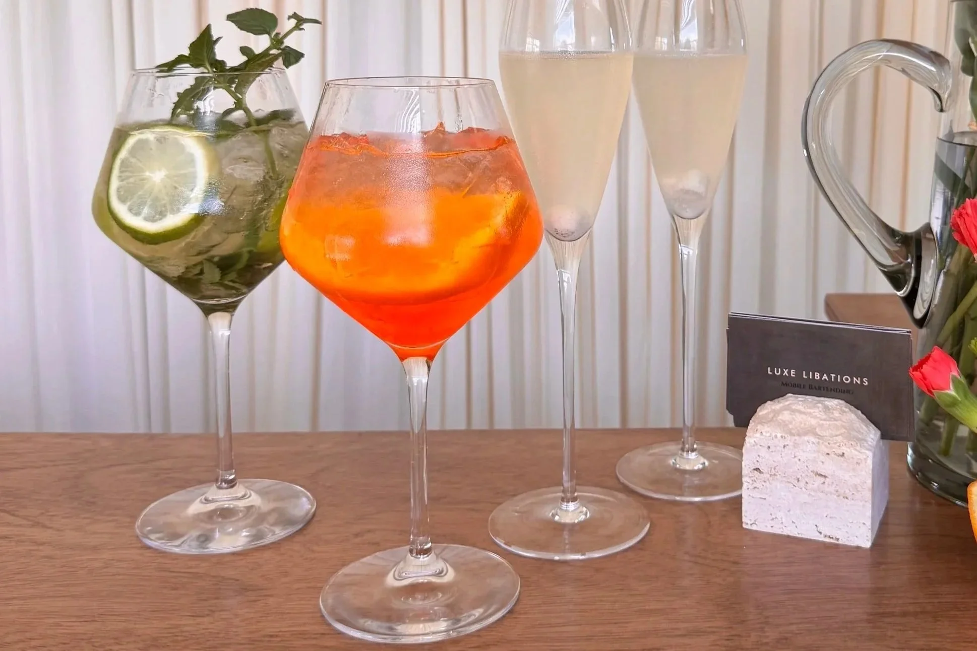 Four elegant glasses of different beverages on a wooden surface, with a potted plant containing lemon slices and mint leaves on the left, and a small black sign card, a stone, and a glass jar with flowers on the right.