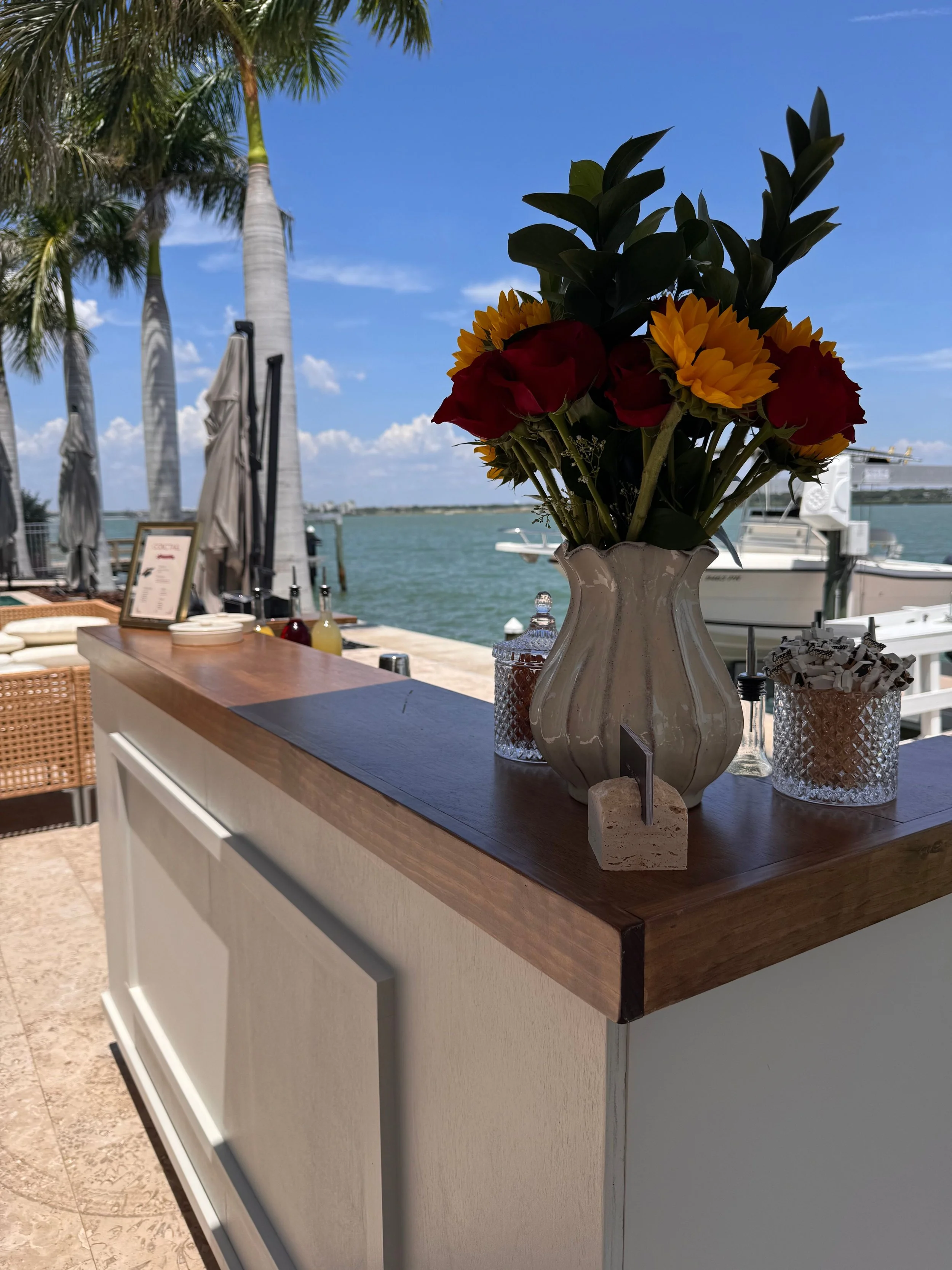 A white ceramic vase with dark green leaves and colorful flowers, including yellow sunflowers and red blooms, on a wooden countertop near a waterfront with boats and palm trees in the background.