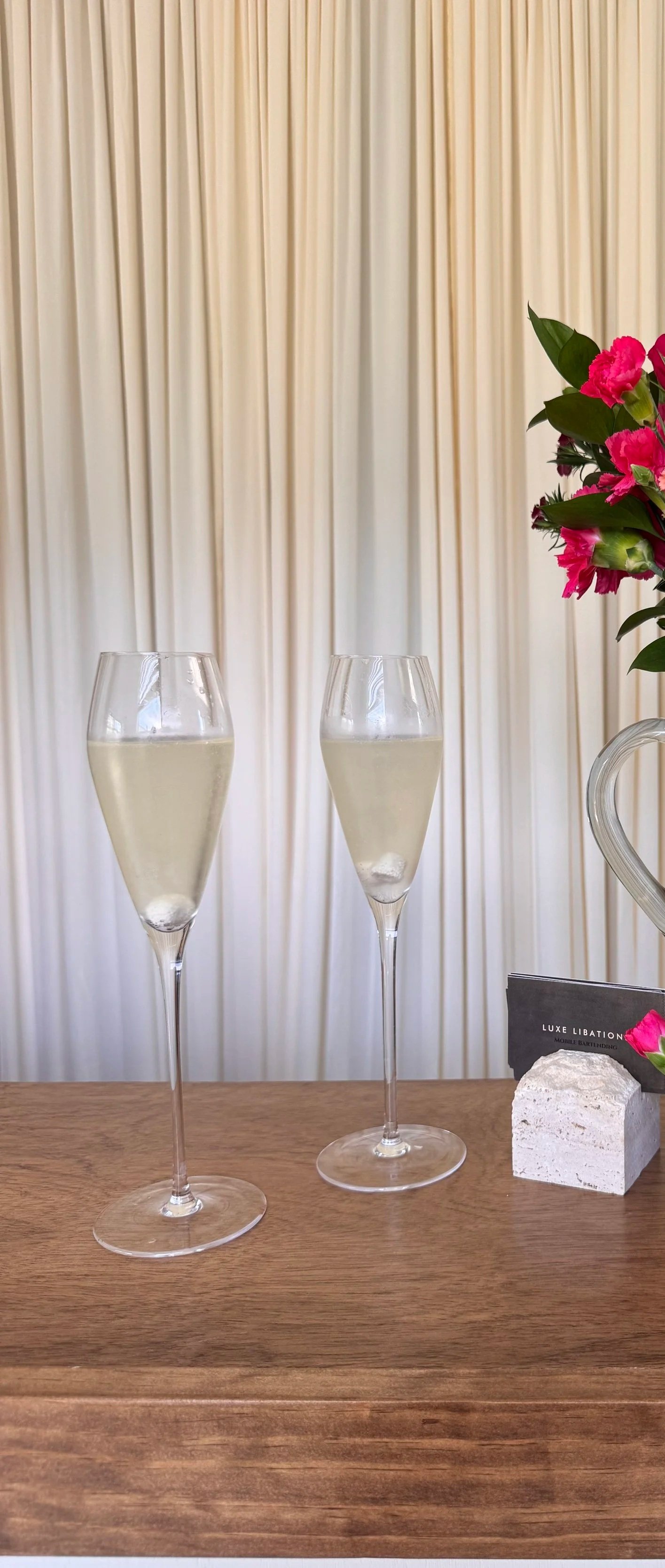 Two glasses of champagne on a wooden table, decorated with a bouquet of pink flowers and a small black sign, against a beige curtain background.