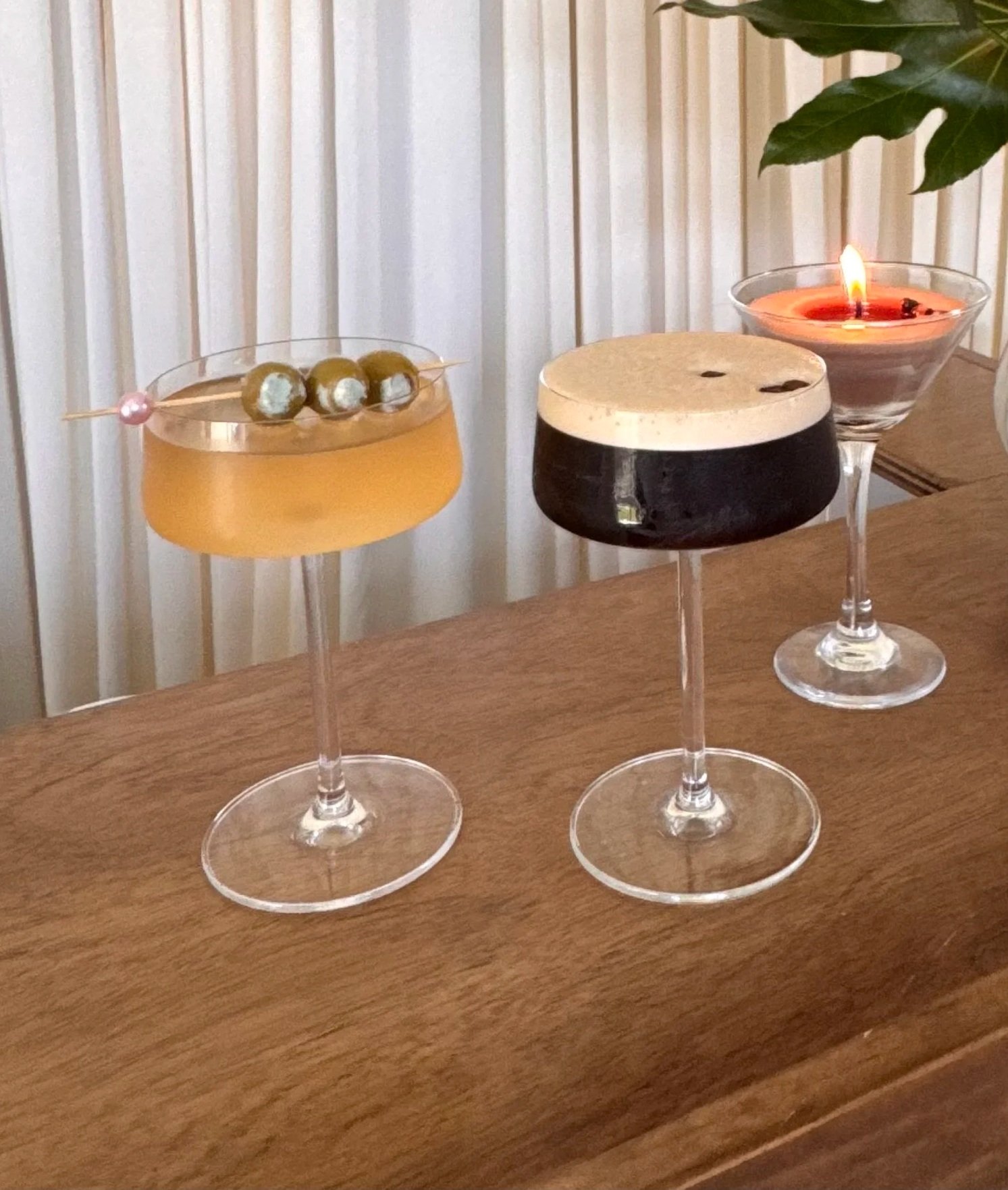 Three cocktails in stemmed glasses, one with green olives and one with an orange beverage and a pink pearl, on a wooden surface with a background of white vertical blinds and a large green leaf.