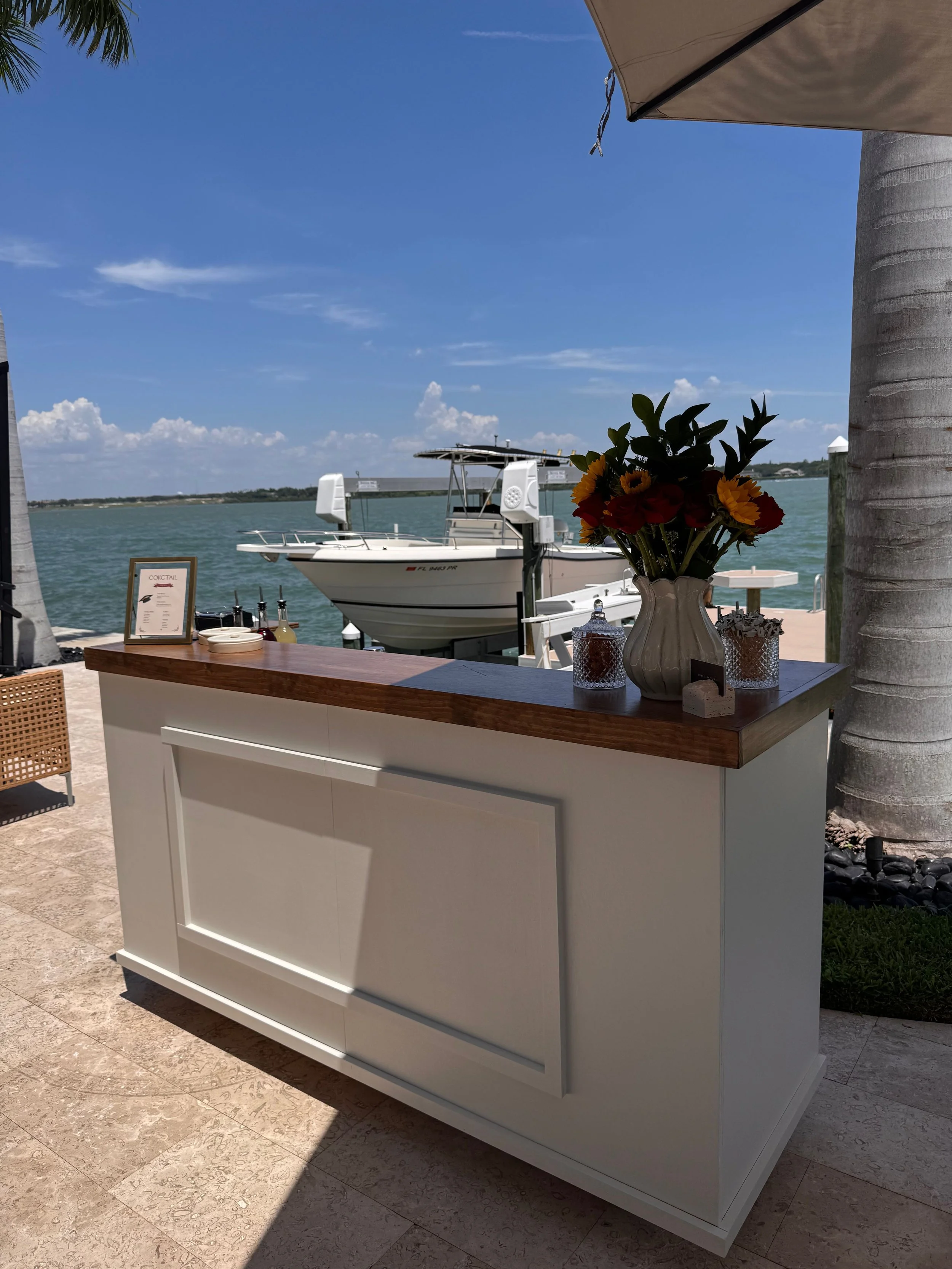 A white outdoor bar with a wooden top, decorated with a vase of colorful flowers, two candle holders, and a small framed cocktail menu, overlooking boats on water under a blue sky.