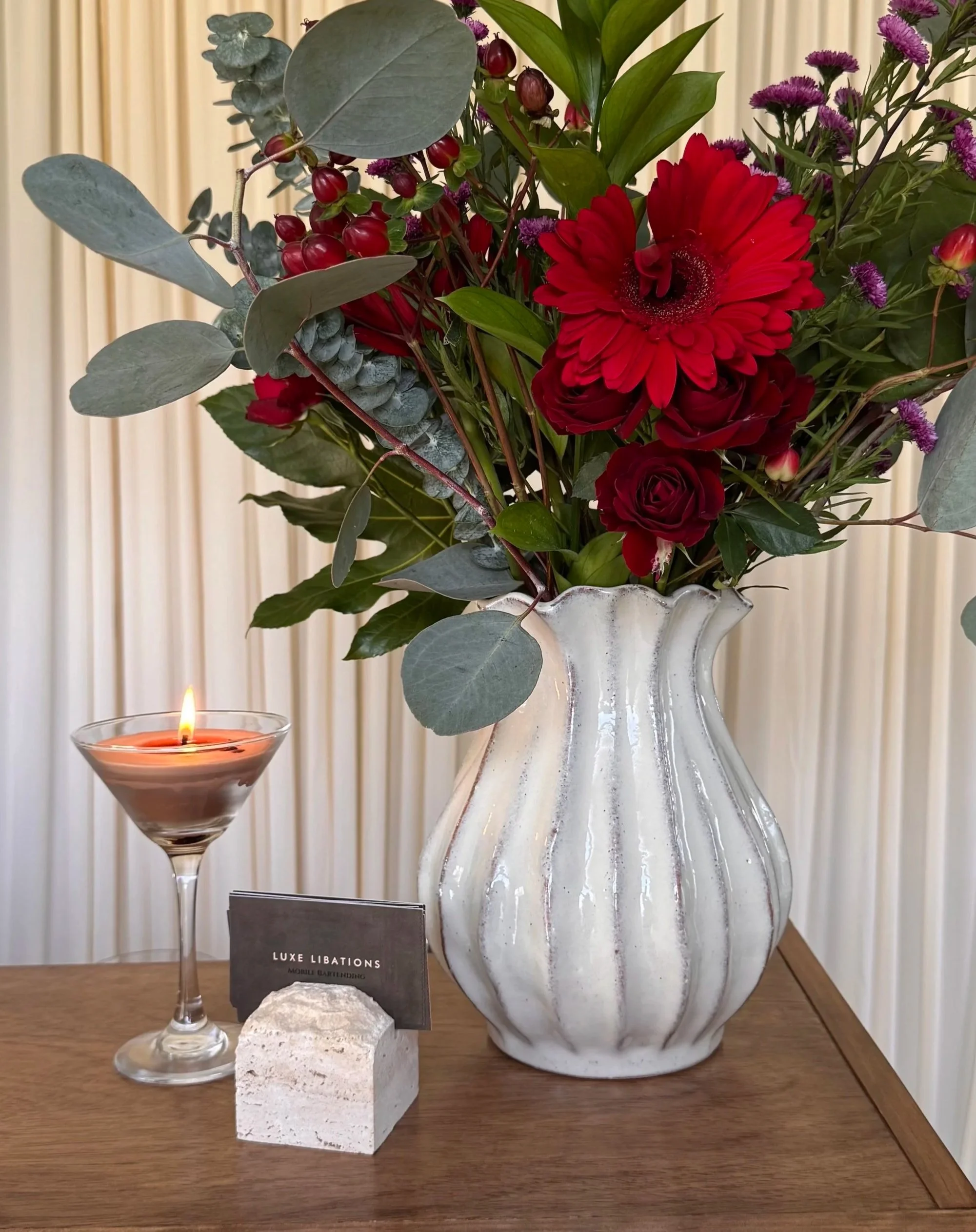 A flower arrangement in a large marble vase with red, purple, and green flowers and leaves. A lit candle in a glass holder is placed in front of the arrangement. There is a small black sign with white text that says 'Luxe Libations' and 'Mobile Bartending'.