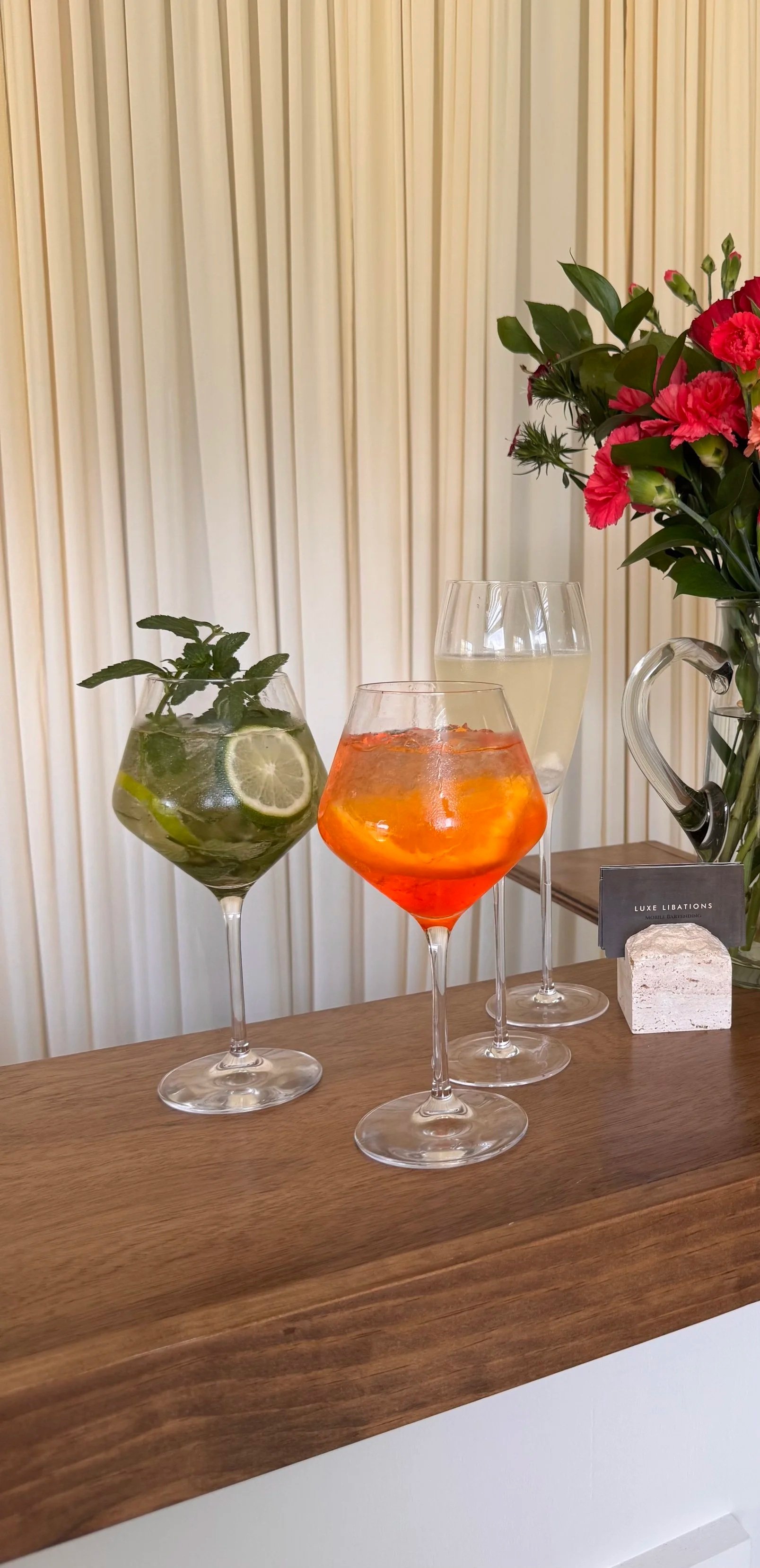 Three colorful cocktails in elegant glasses on a wooden table, with a vase of pink flowers and a small sign in the background.