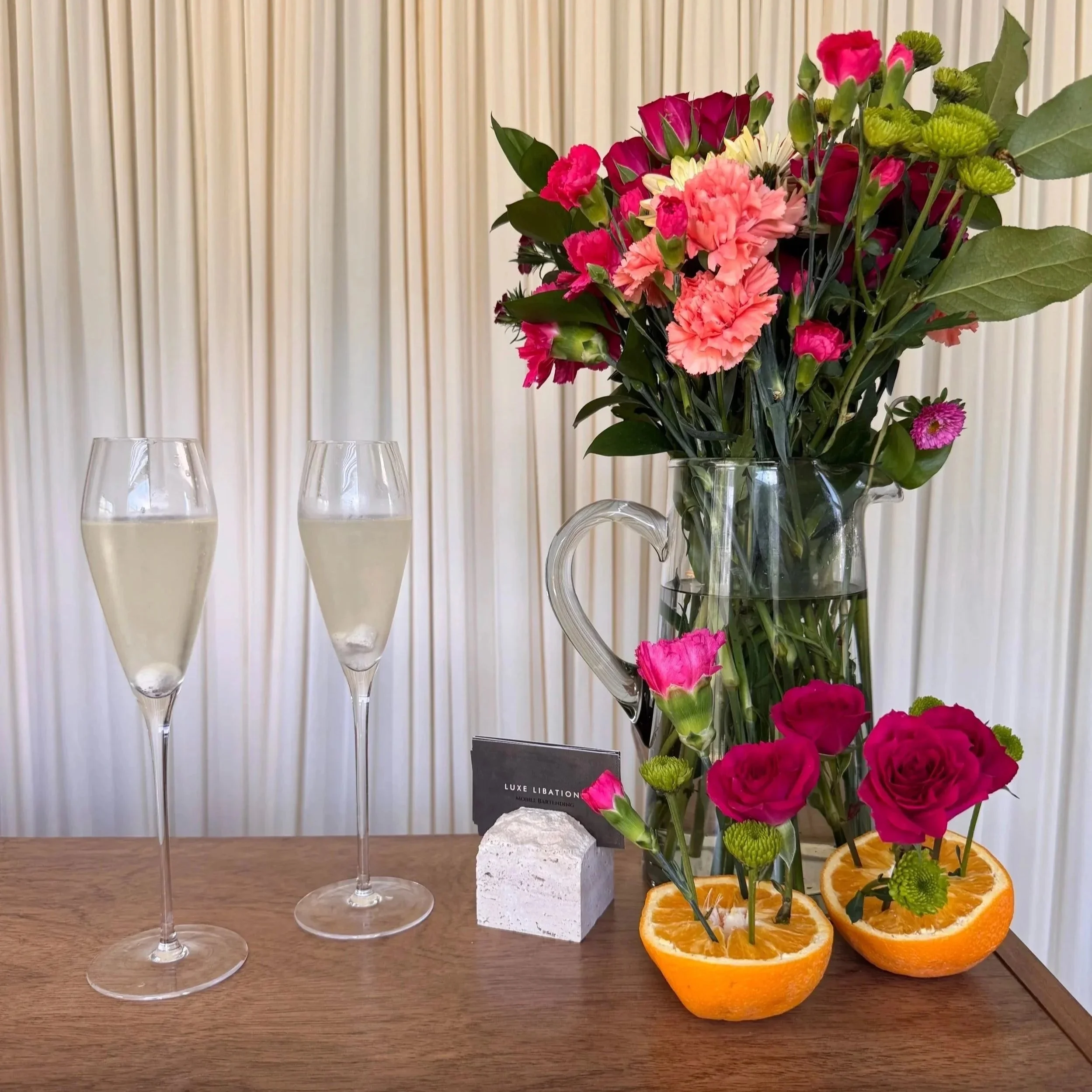Three empty wine glasses, a white stone with a black label, a glass pitcher with a bouquet of pink and red flowers, and slices of oranges with garnish on a wooden table.