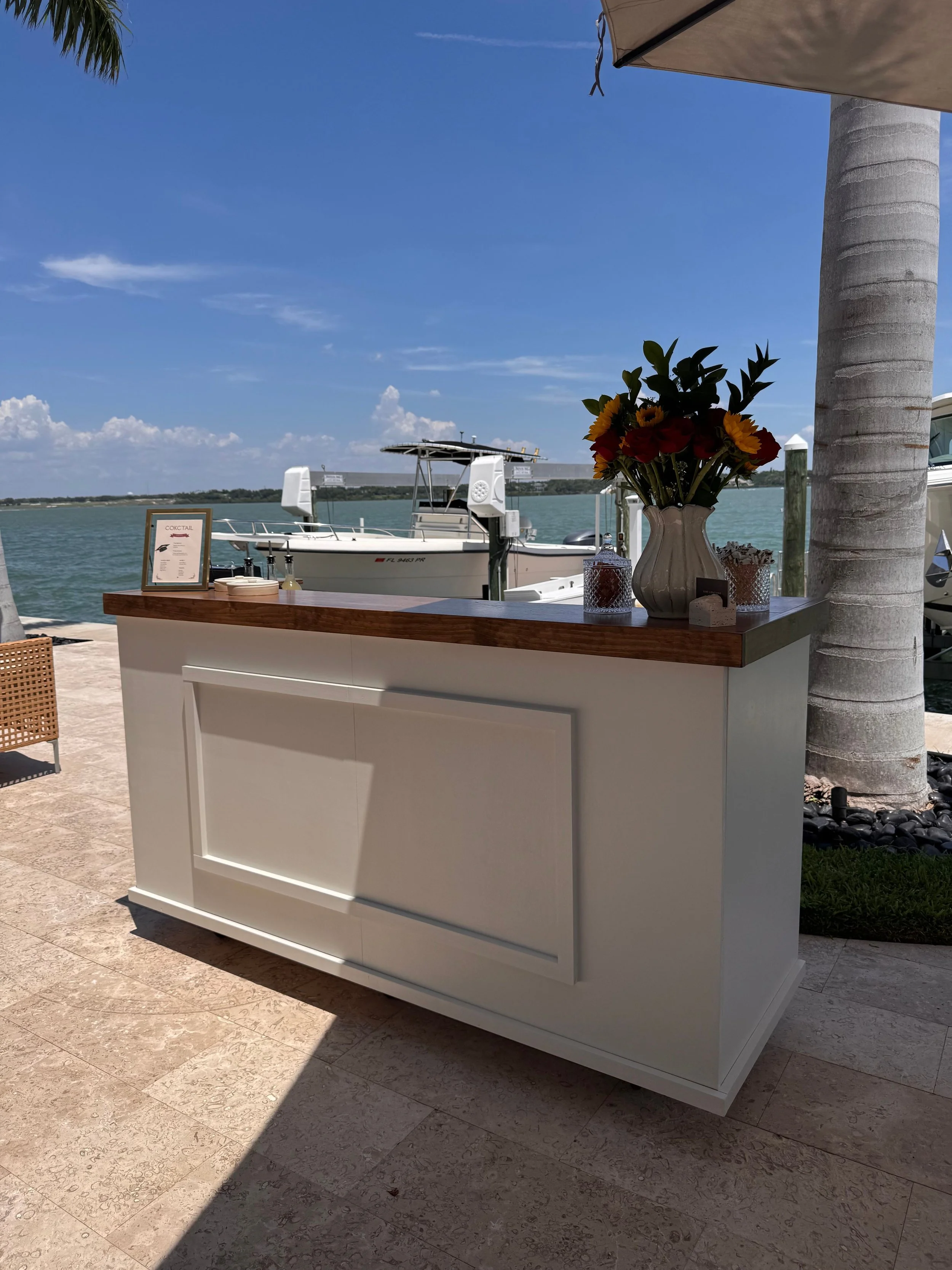 A white outdoor bar or counter with a wood top, decorated with a vase of flowers and candles, overlooking a body of water with boats and a blue sky.
