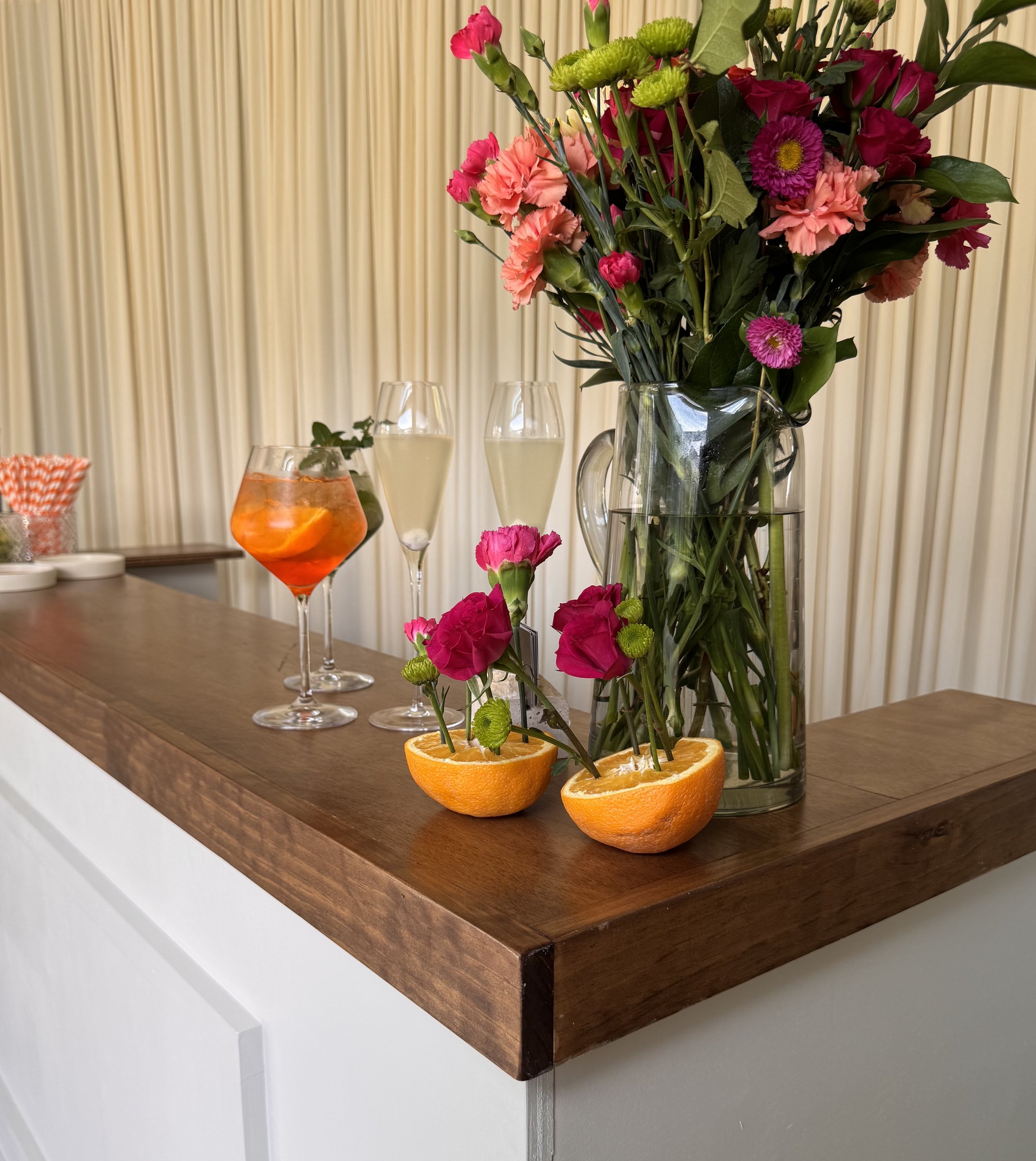 A glass vase with a large bouquet of pink, purple, and green flowers on a wooden table, with sliced oranges used as a base. Two champagne flutes are behind the vase, and a drink with orange slices is also on the table. The background is a beige curtain.