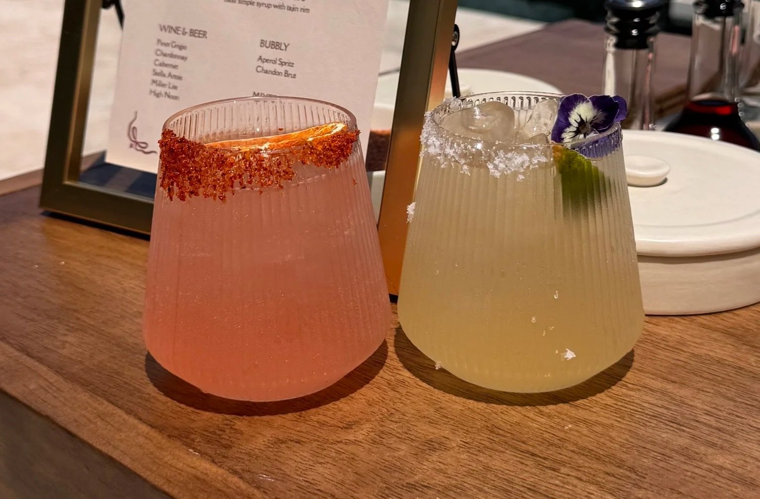 Two colorful cocktails on a wooden table, one pink with a chili salt rim and a slice of orange, the other pale yellow with a sugar rim, a lemon slice, and a purple flower garnish.