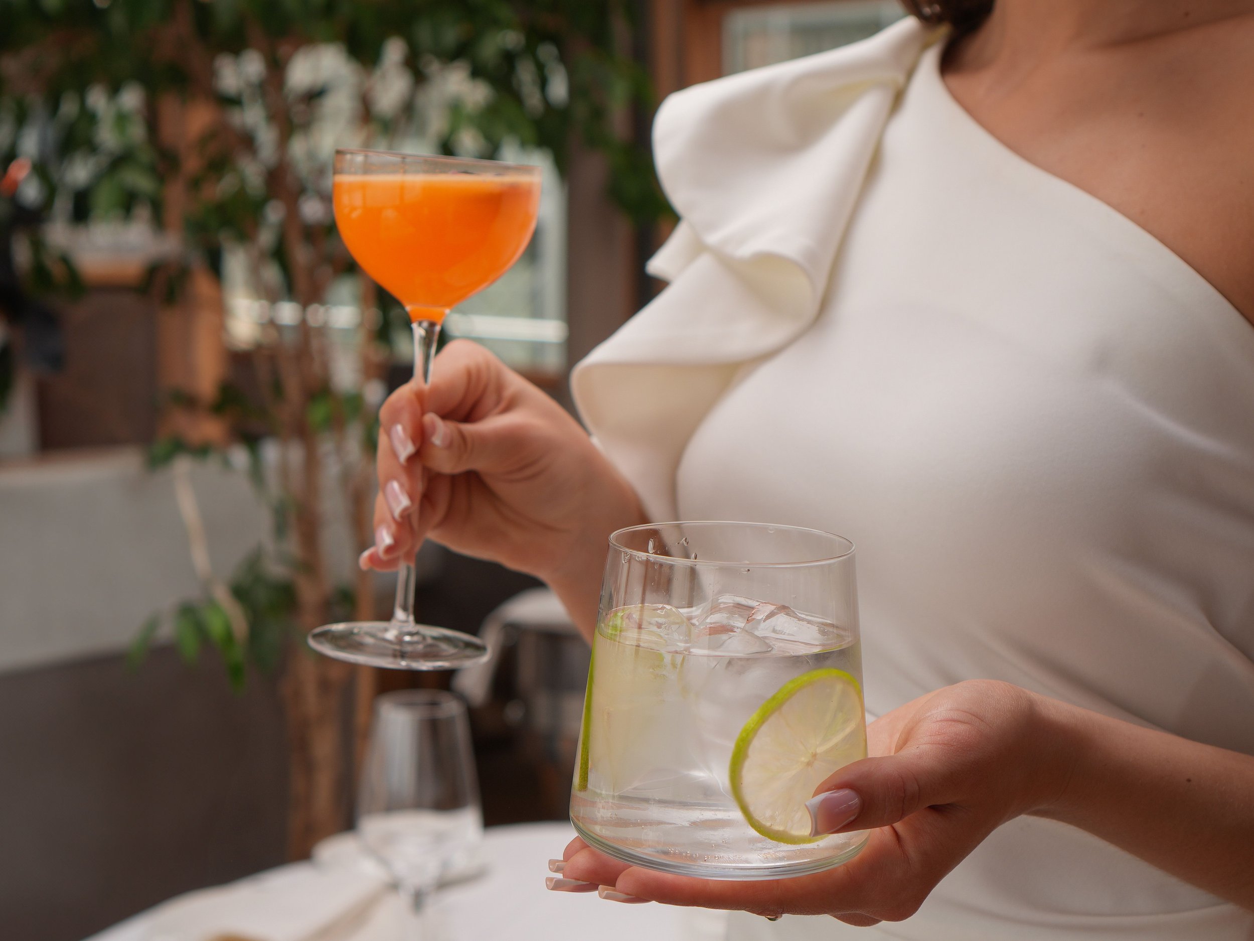 A woman in a white outfit holding a glass of cocktail with lemon and lime slices, with other drinks visible on the table and a blurred background of plants and windows.