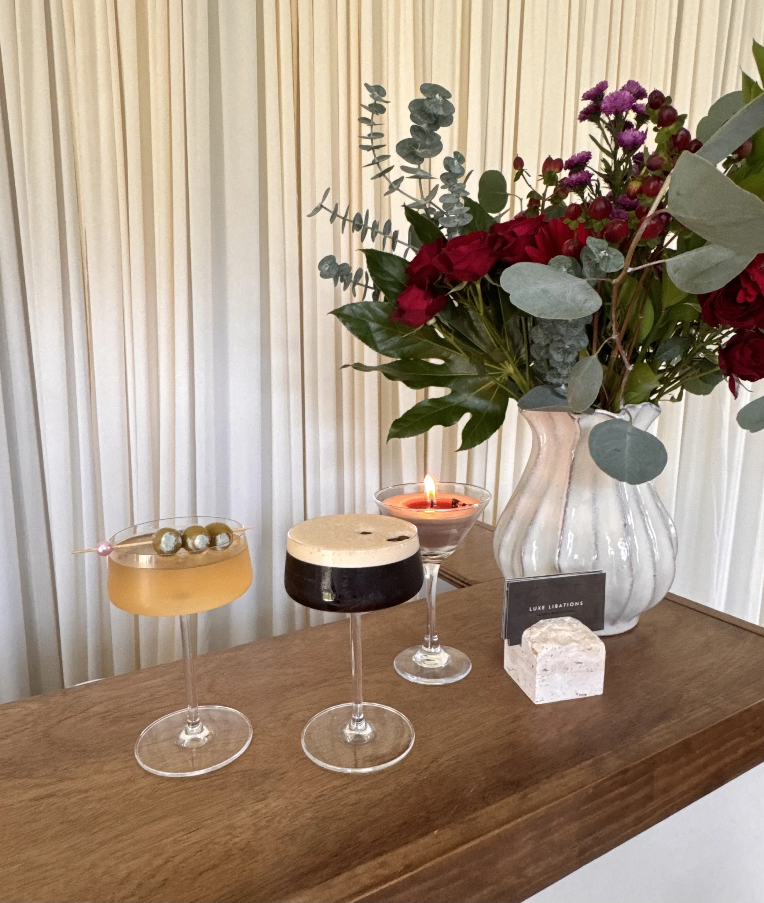 Display table with three elegant champagne flutes, a candle, and a decorative stone. In the background, a large flower arrangement with green leaves, red and purple flowers, and eucalyptus in a clear glass vase.