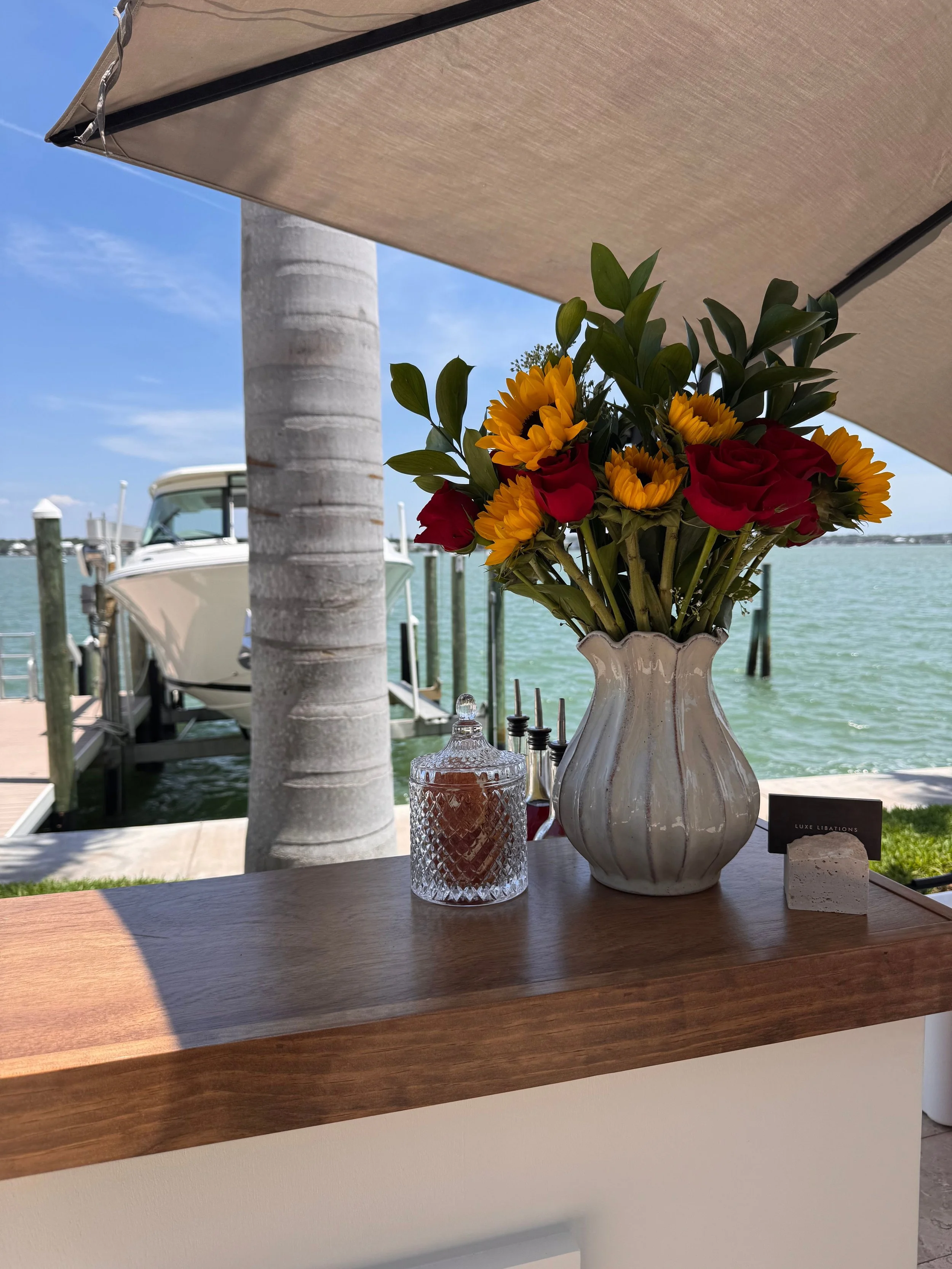 A table with a vase containing sunflowers, red roses, and greenery, a glass jar with a candle inside, and a small sign with the text 'Luxe Libations' on a waterfront with boats and a palm tree in the background. A beige patio umbrella provides shade.