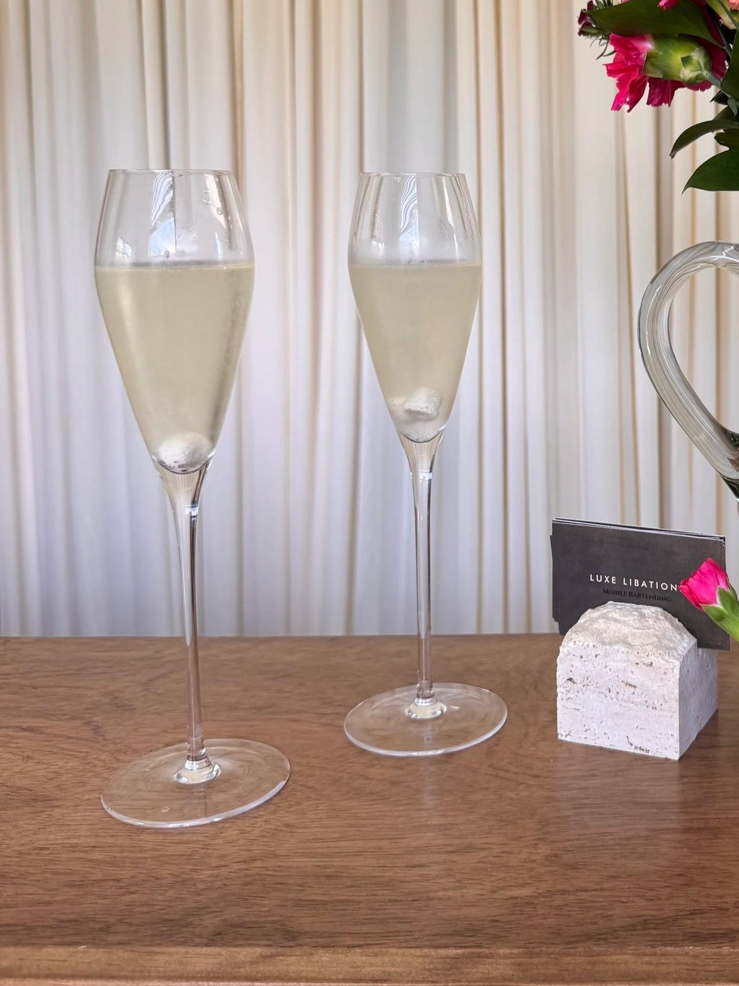 Two champagne flutes filled with sparkling wine, a small bouquet of pink flowers in a glass vase, and a card that reads 'LUXE LIBATION MODEL BAKELDING' on a wooden surface with a light-colored background.