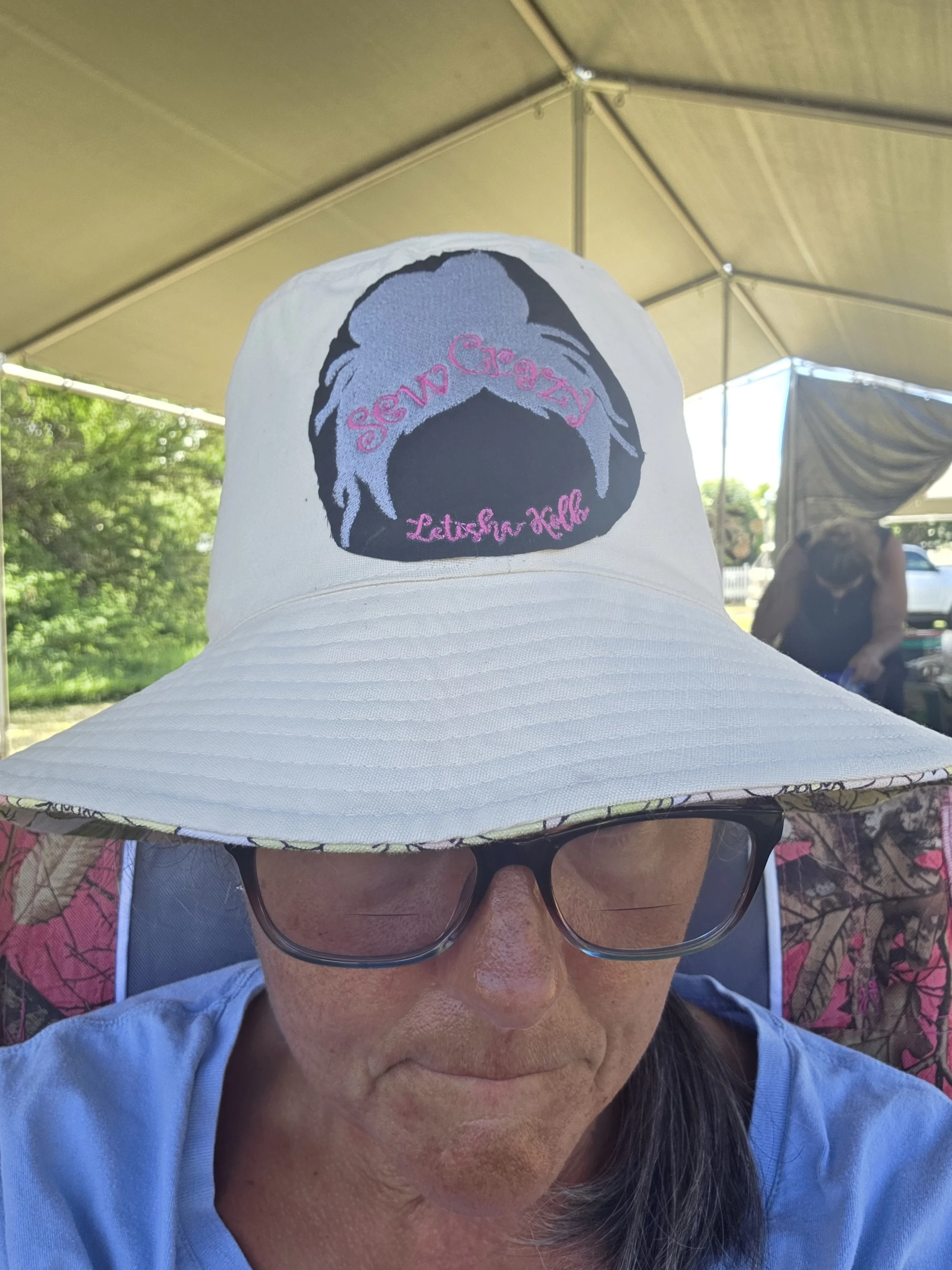 A woman wearing a large, white sunhat with an embroidered design and the words "Stay Crazy Let Her Roll" on it, and sunglasses, covers her face with her head tilted downward.