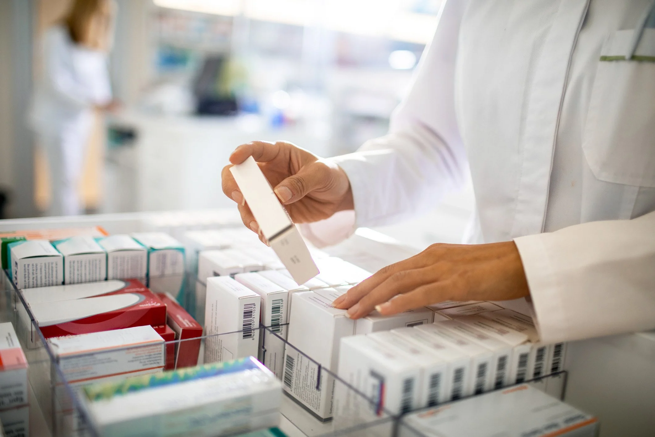 A person in a white coat browsing shelves of medicine in a pharmacy.