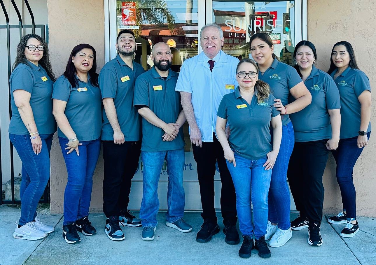 Group of nine pharmacy staff members standing outside Sunrise Pharmacy store, smiling for a photo. They are wearing matching blue shirts with Sunrise Pharmacy logo and nametags.