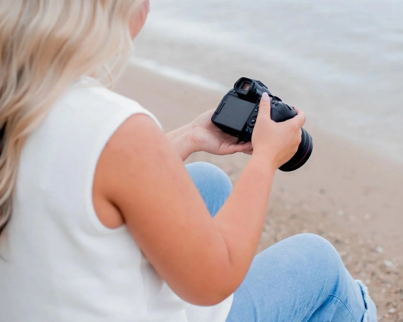A woman sitting on a beach shore holding a camera and looking at the camera's screen.