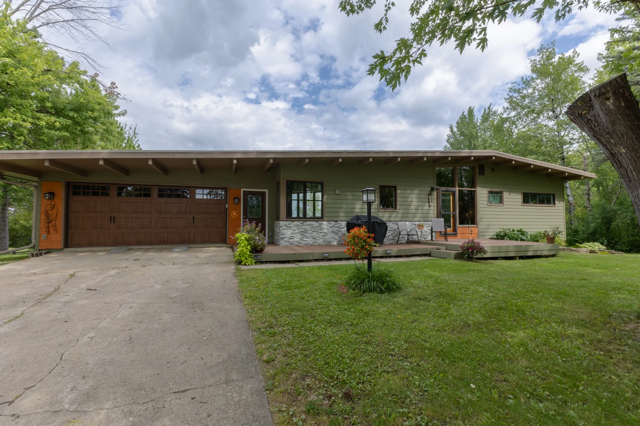 A single-story house with a green exterior, stone accents, and a wooden garage door in a lush yard with trees and flowers, under a partly cloudy sky.