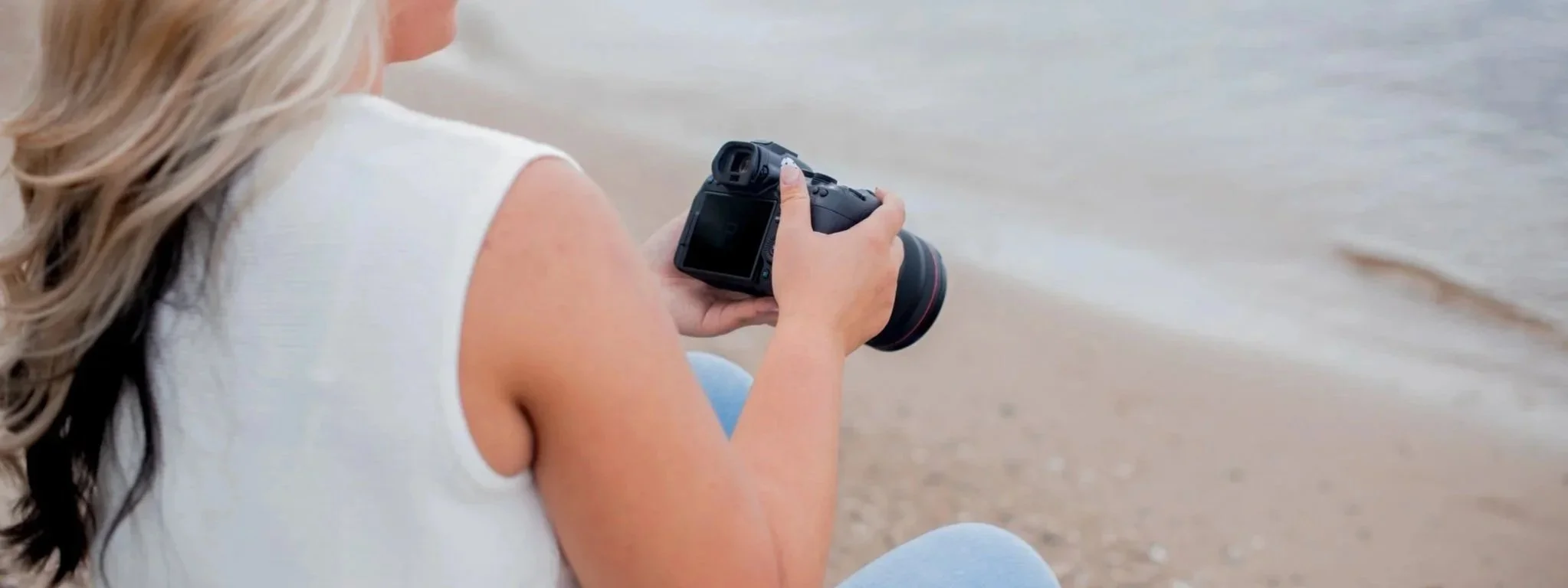 Person holding a DSLR camera while sitting on a beach, with the ocean in the background.
