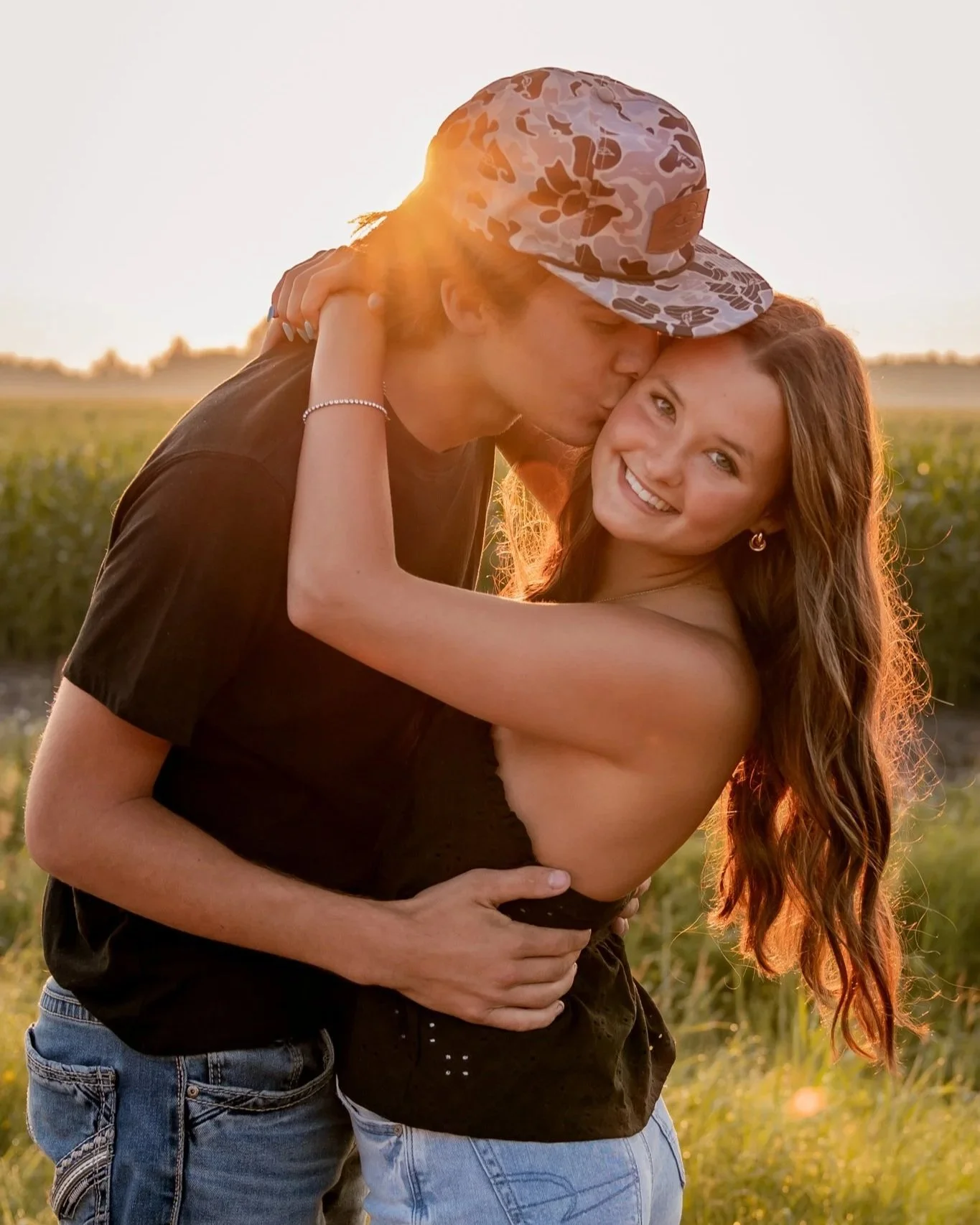 A young couple outdoors during sunset, with the man wearing a camouflage cap and the woman with long, wavy hair, smiling as he kisses her on the forehead. They are embracing in a field.