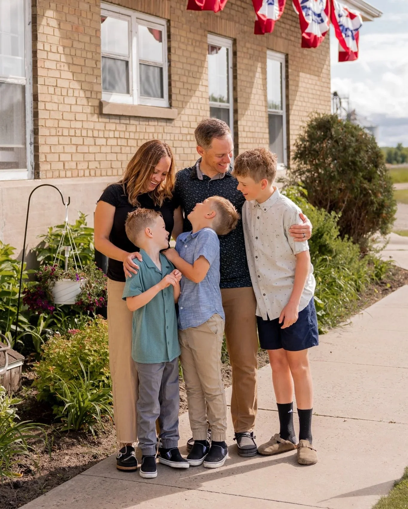 A family of five standing outside on a sidewalk near a brick house, smiling and laughing together. The family includes two adult parents and three young boys. The house is decorated with red, white, and blue bunting, indicating a patriotic celebration.