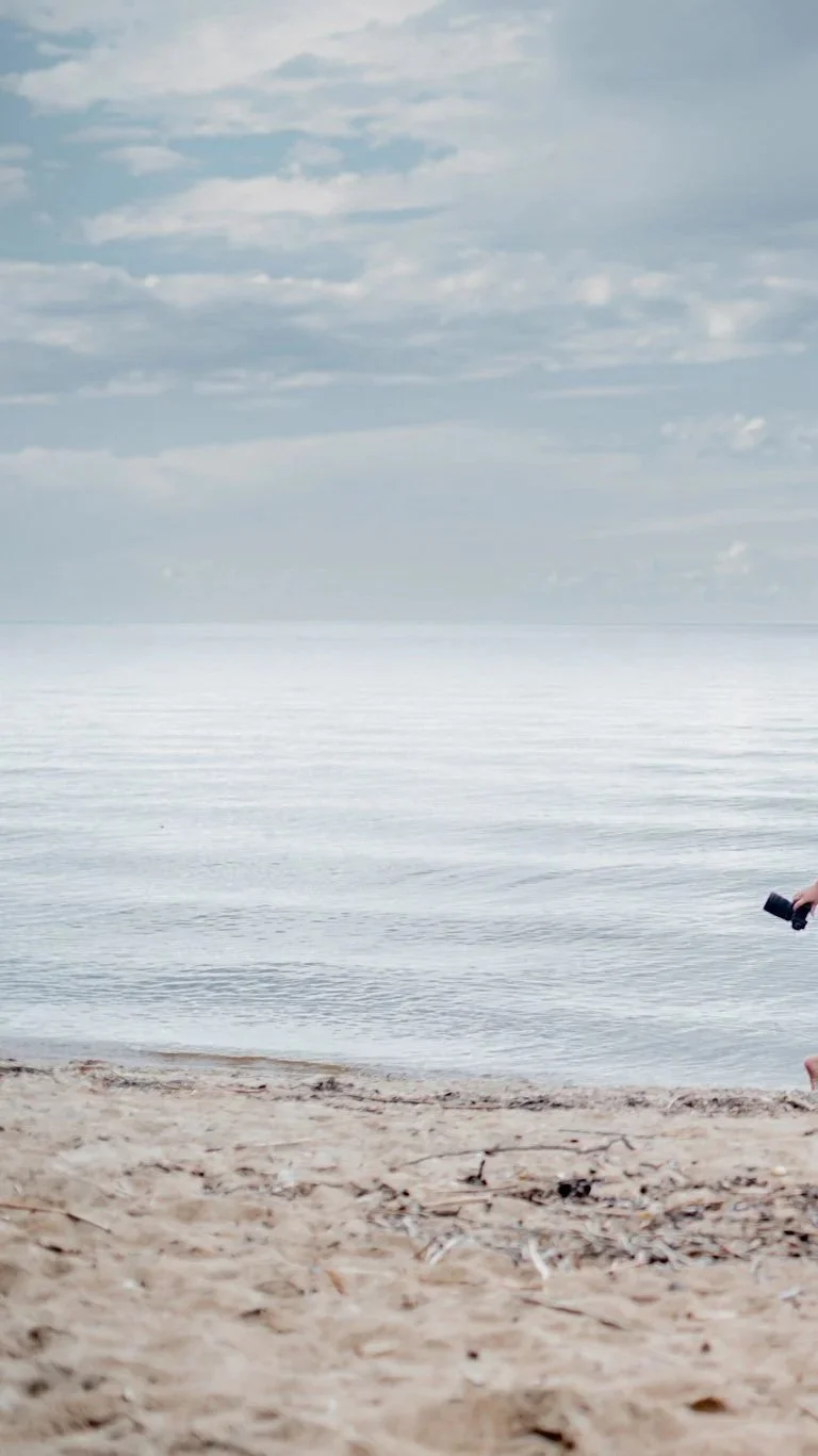 A person is sitting on a sandy beach holding binoculars, facing the calm ocean with a cloudy sky overhead.
