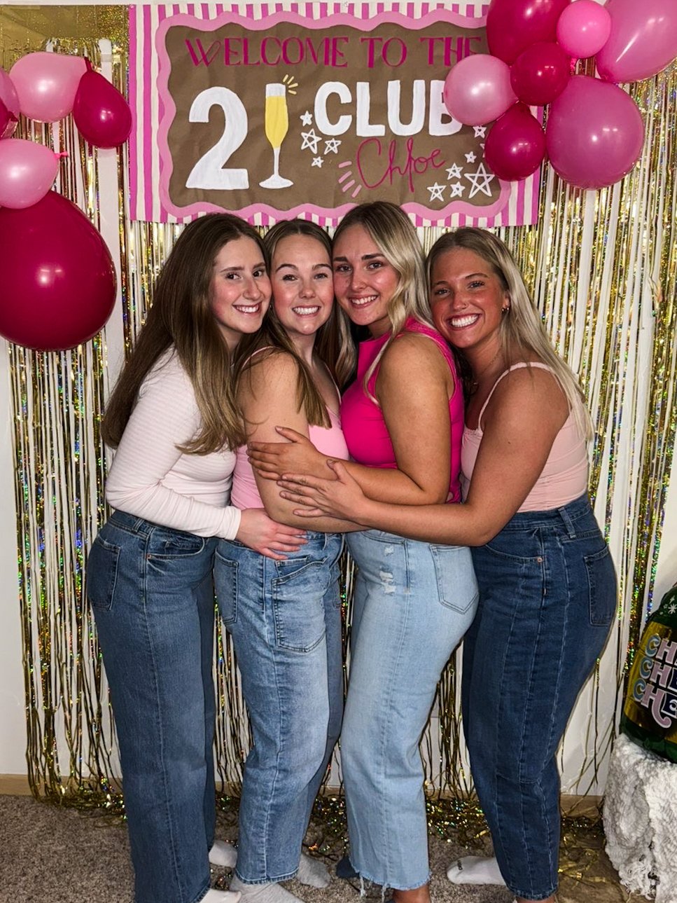 Four women smiling and hugging in front of a pink, brown, and white sign that says "Welcome to the 21 Club Chloe," with pink and burgundy balloons and gold tinsel curtains behind them.