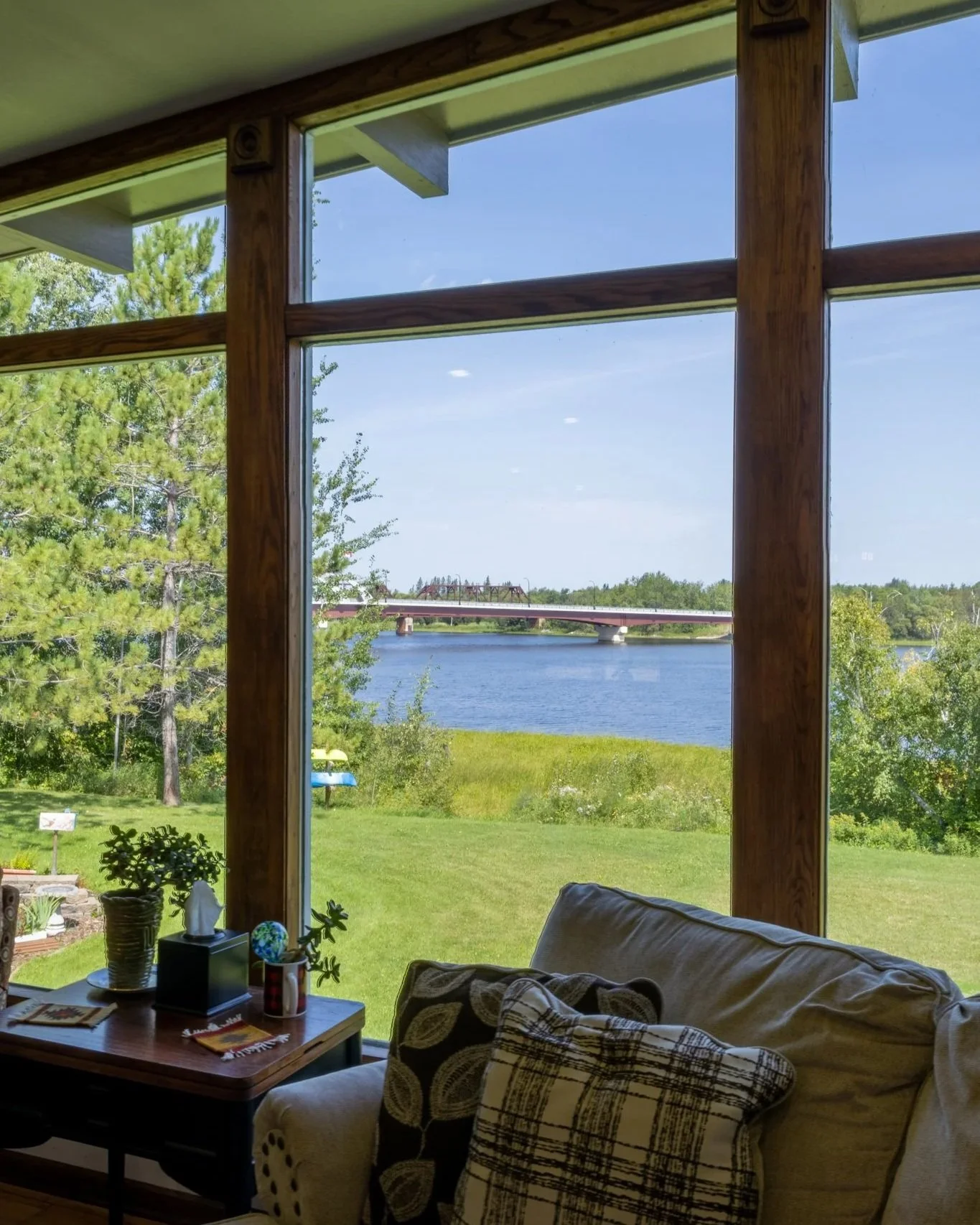 Living room with large window looking out over a river and bridge, with a sofa, pillows, and a small table with decorative items inside.