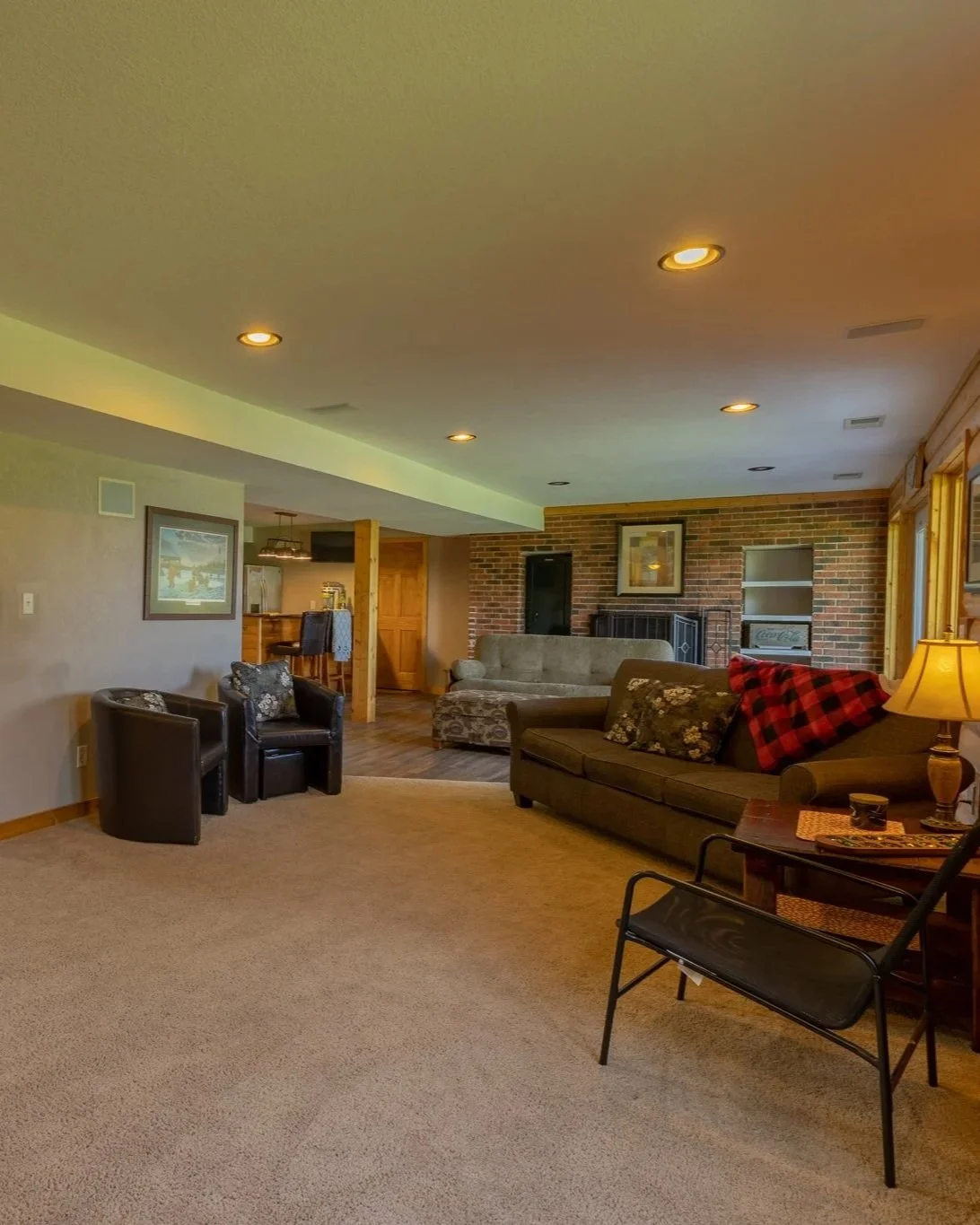 Living room with beige carpet, brown sofa with plaid blanket, black armchairs, brick fireplace, framed artwork, wooden shelves, and window with curtains.