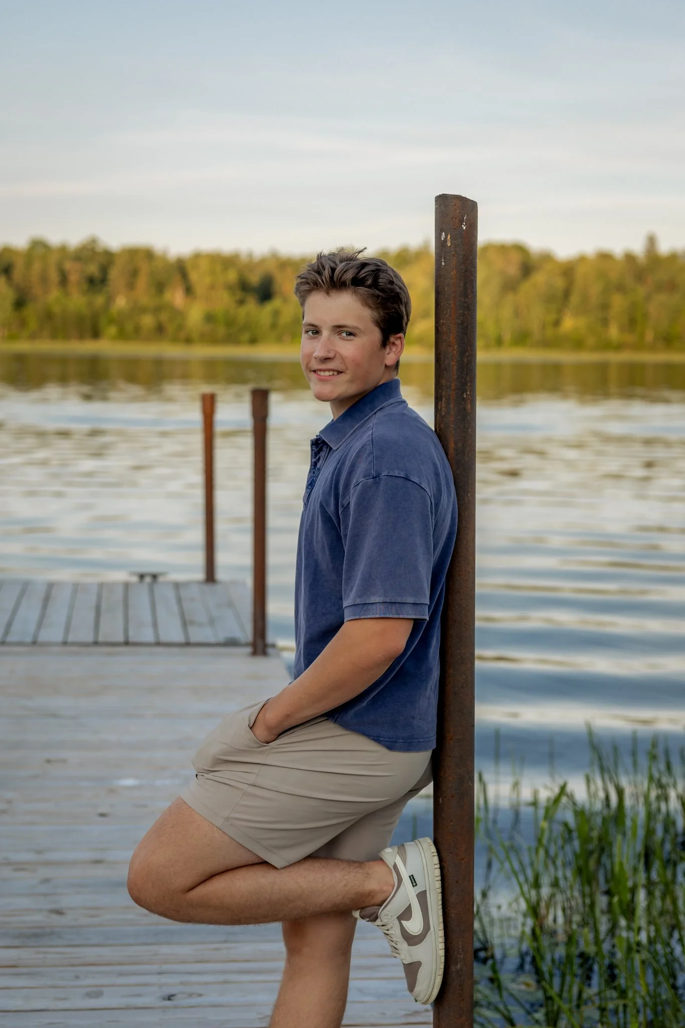A young man with short brown hair, wearing a blue polo shirt and beige shorts, leaning against a rusty metal post on a dock by a lake. The background shows a calm lake, green trees, and a clear sky.