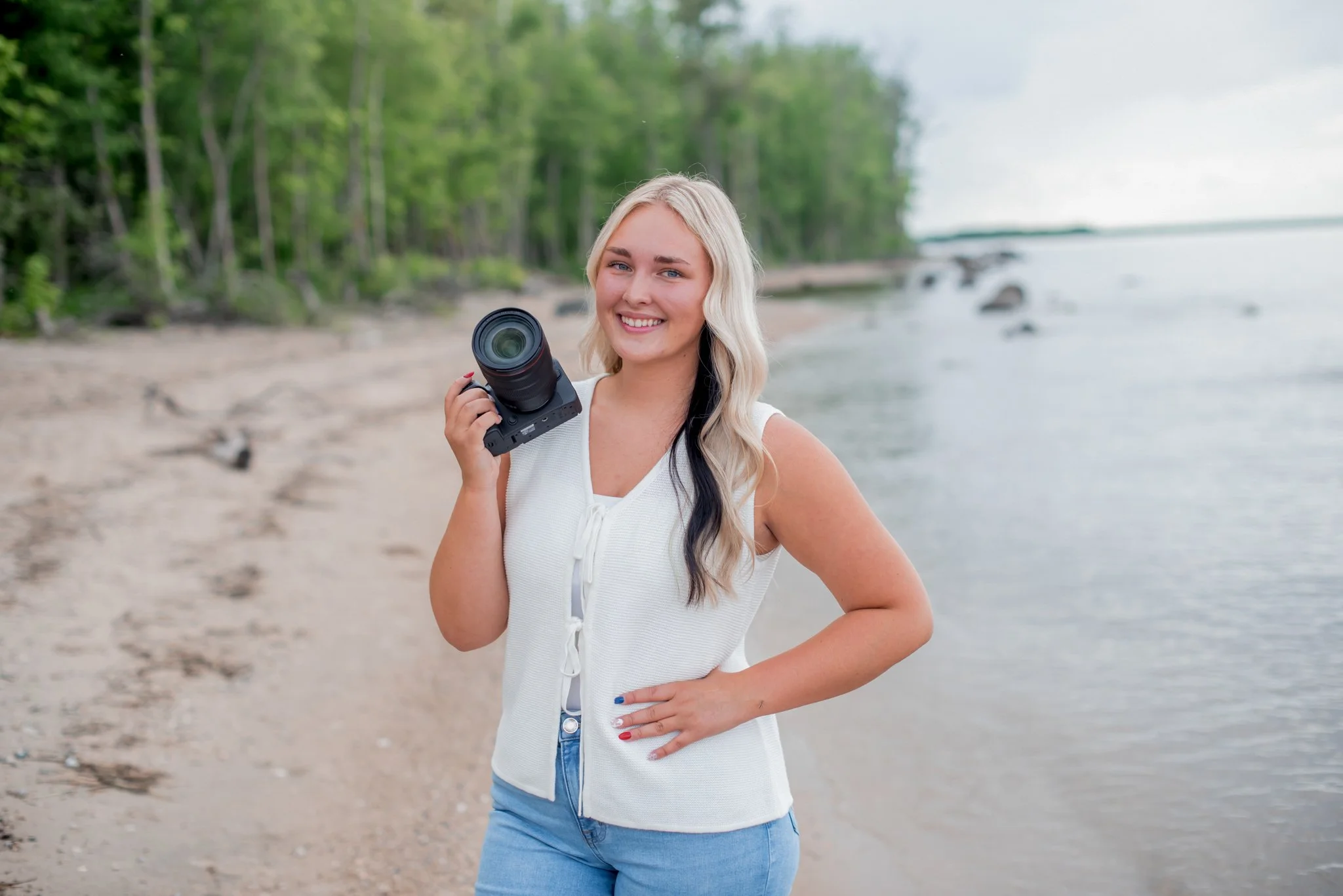 A young woman with blonde hair, wearing a white sleeveless top and blue jeans, smiling and holding a camera on a beach with green trees and water in the background.