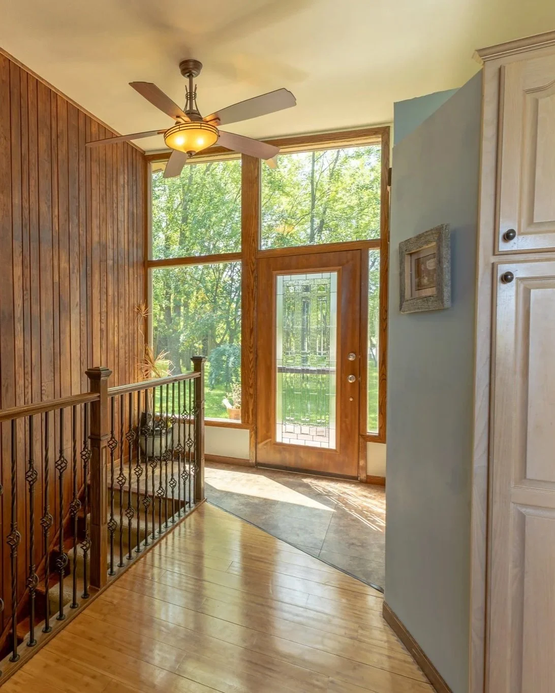 Bright entryway with wooden door and large glass windows, wooden floors, ceiling fan, and railing along stairs, with lush green outdoor scenery visible through the windows.