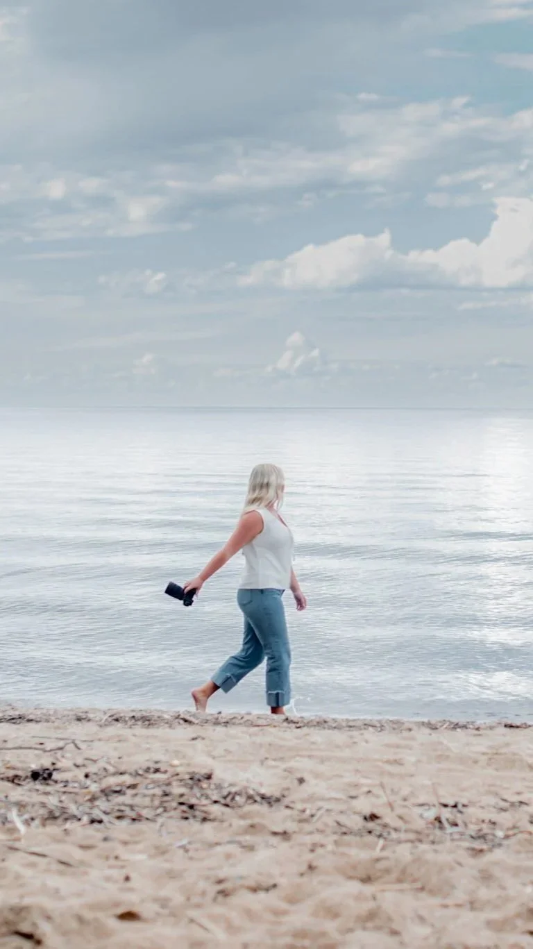A woman with blonde hair in a white sleeveless top and blue jeans walking barefoot along the sandy shore, holding binoculars in her right hand, with calm water and cloudy sky in the background.