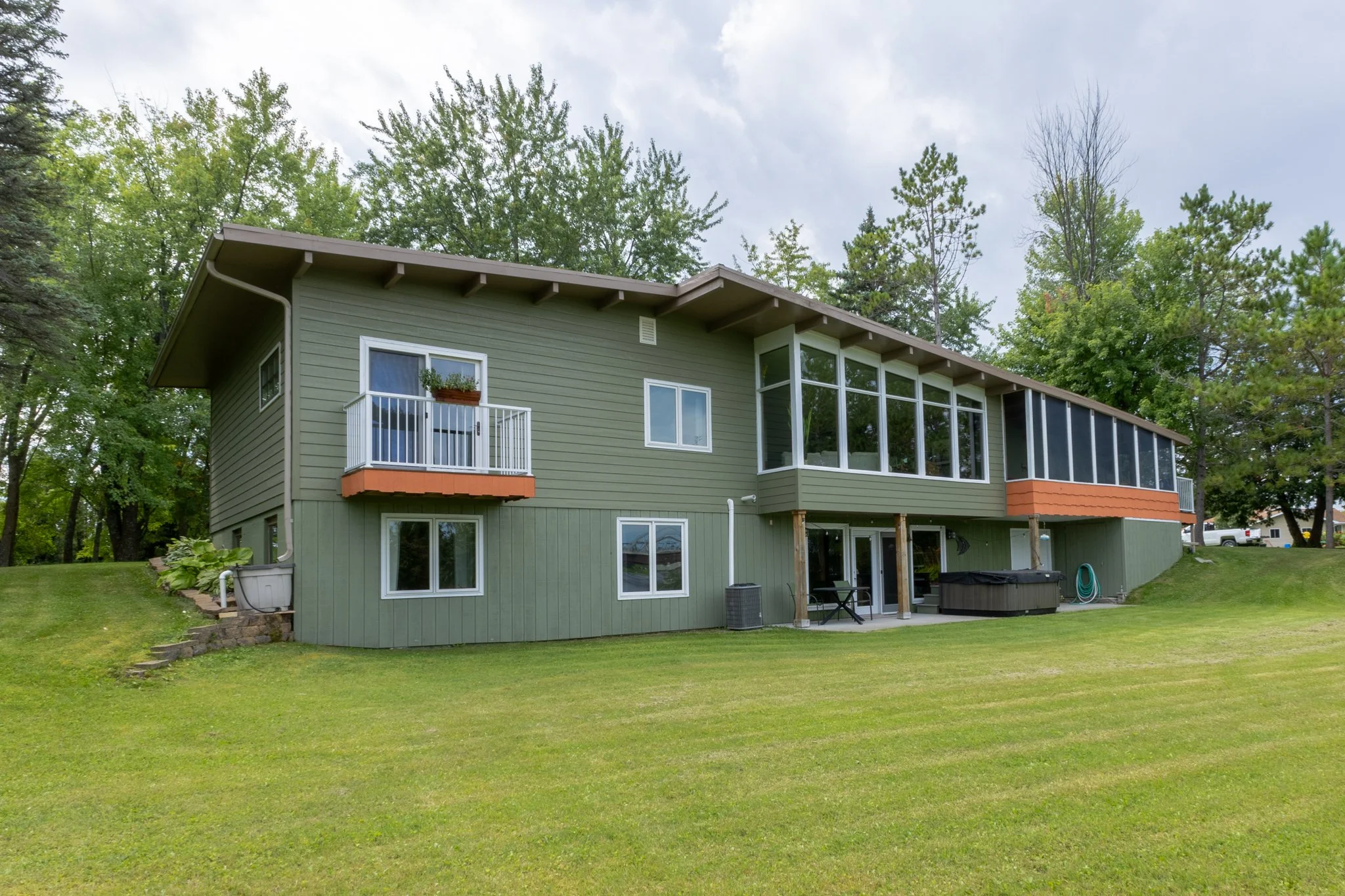 The back view of a green, two-story house with large windows and a small balcony, surrounded by a lush green lawn and tall trees, with a cloudy sky above.