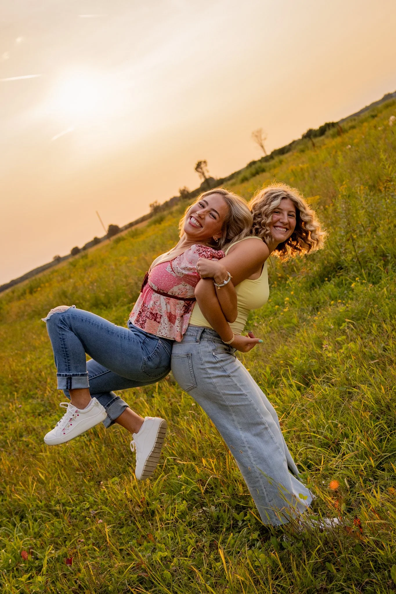 Two women smiling and hugging in a grassy field with a sunset sky, one woman is lifted by the other, both wearing casual clothes.