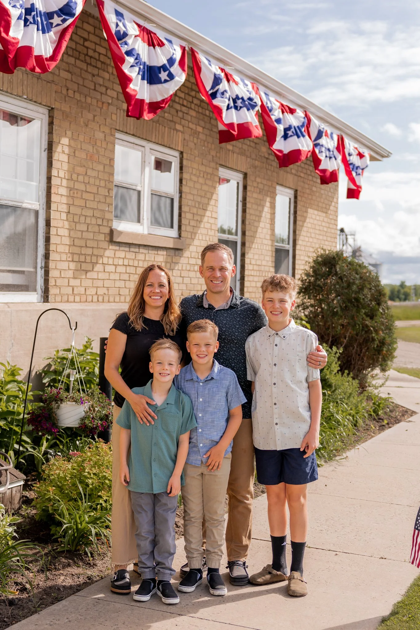 Family of five standing on a sidewalk in front of a house decorated with red, white, and blue patriotic bunting, with American flags on the porch, during daytime with partly cloudy sky.