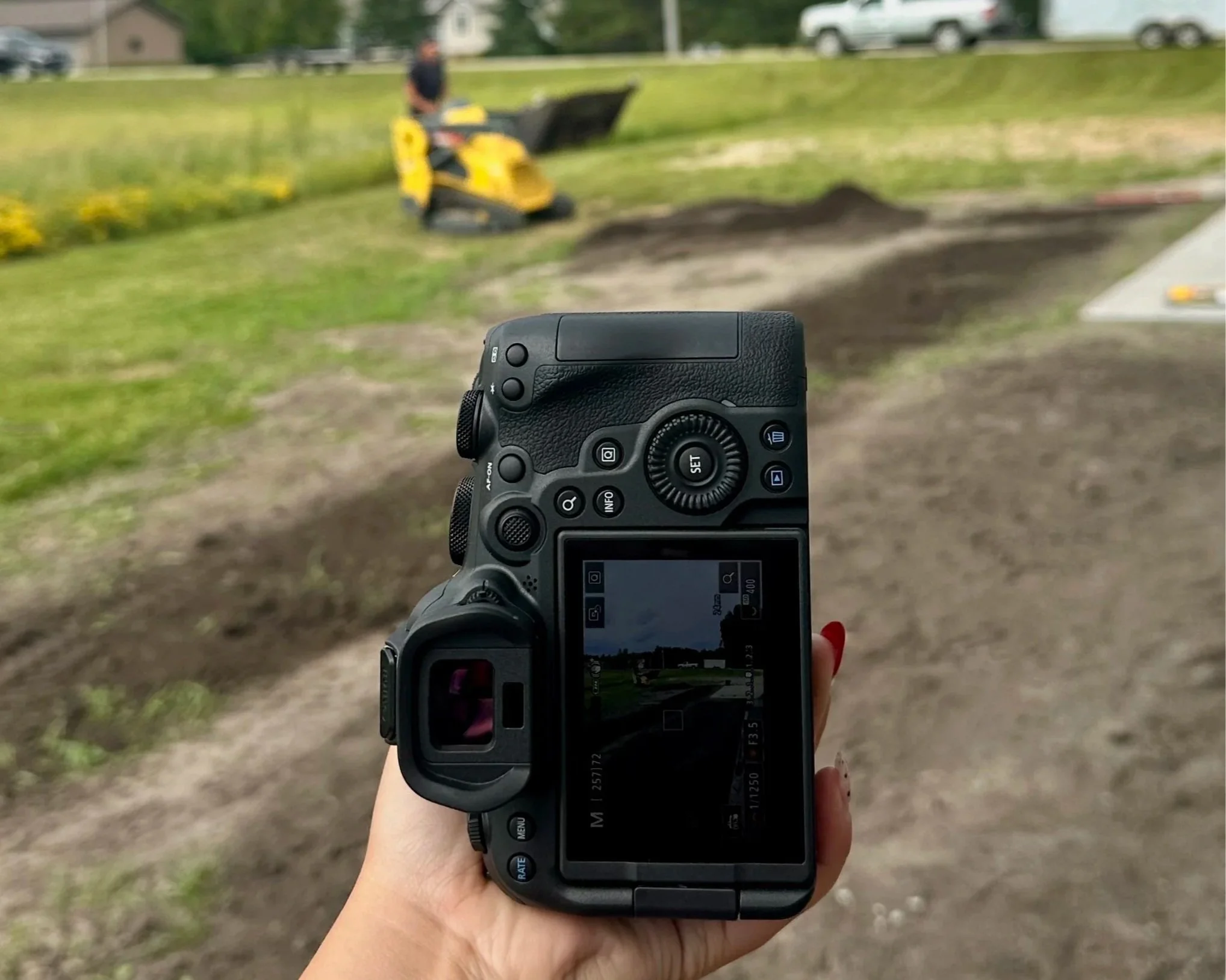 A person holding a camera points towards a blurred view of a person operating a yellow compactor on a grassy outdoor area, with a sidewalk and parked cars in the background.